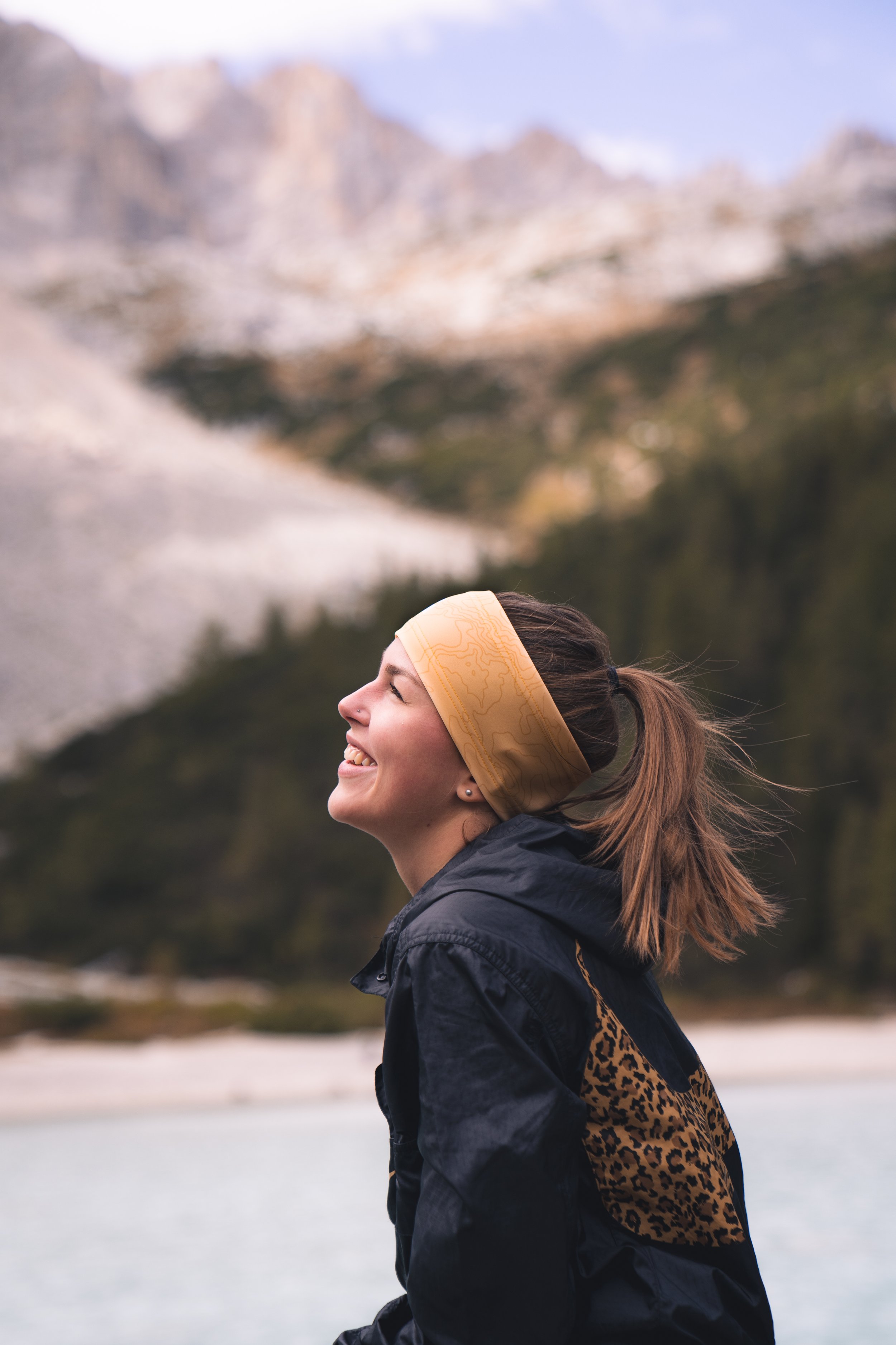 A woman smiling outdoors with mountains and trees in the background, wearing a yellow headband and a black jacket with leopard print accents.