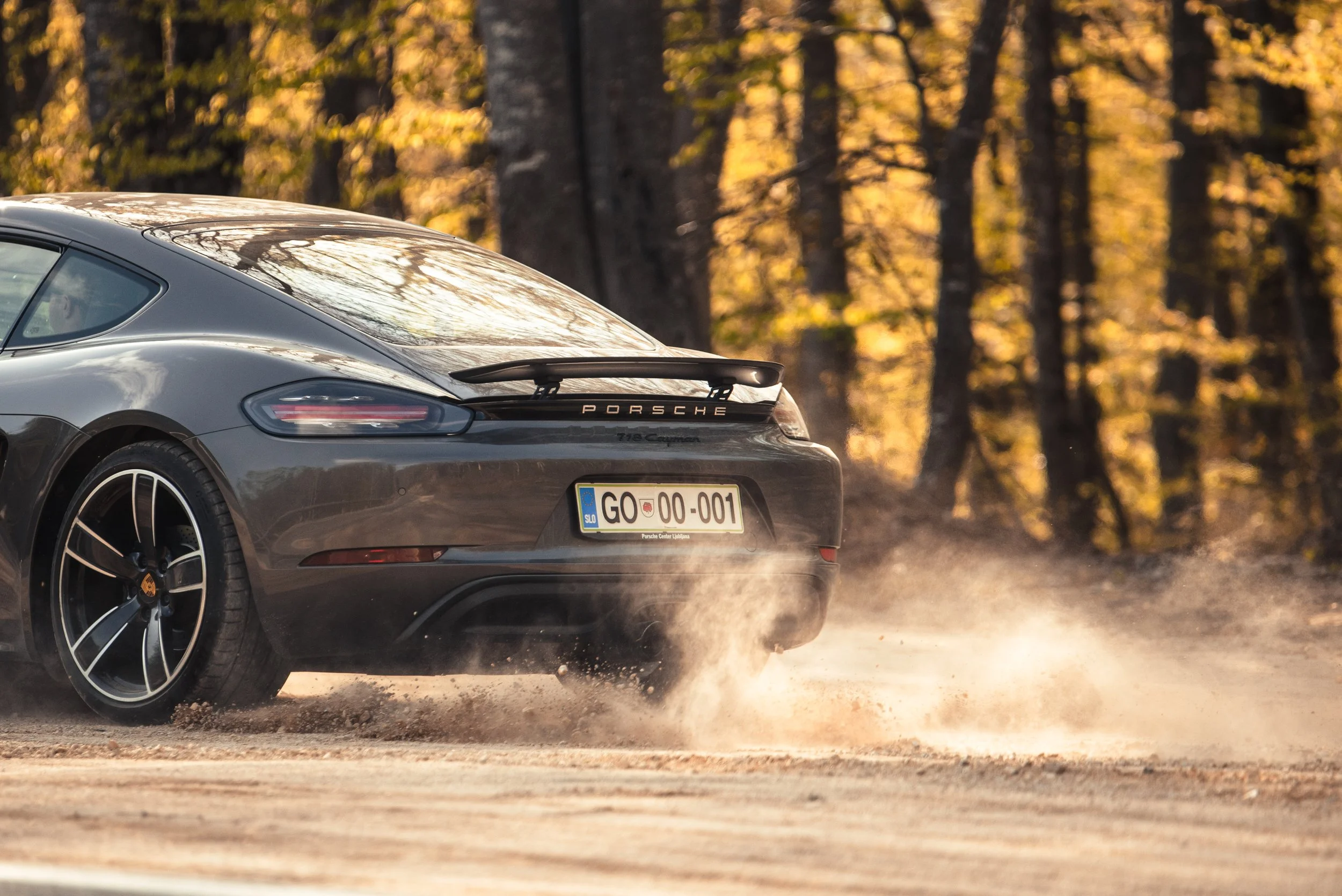 A gray Porsche 718 Cayman driving on a dirt road in a forest with autumn-colored trees, kicking up dust behind it.