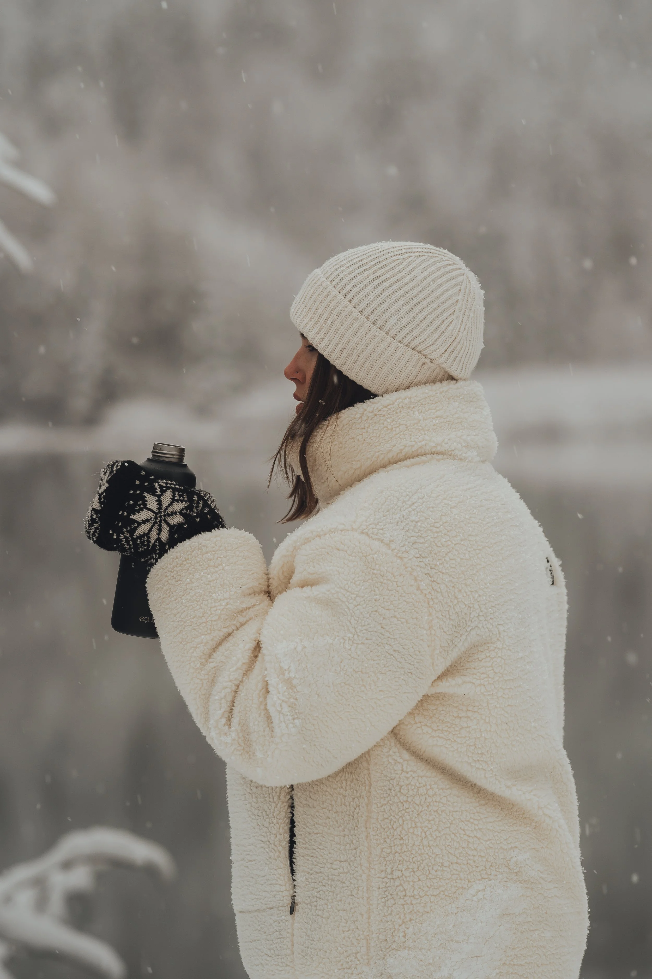 A woman dressed warmly in a cream fleece jacket, knit hat, and gloves, holding a travel mug outdoors in a snowy landscape.