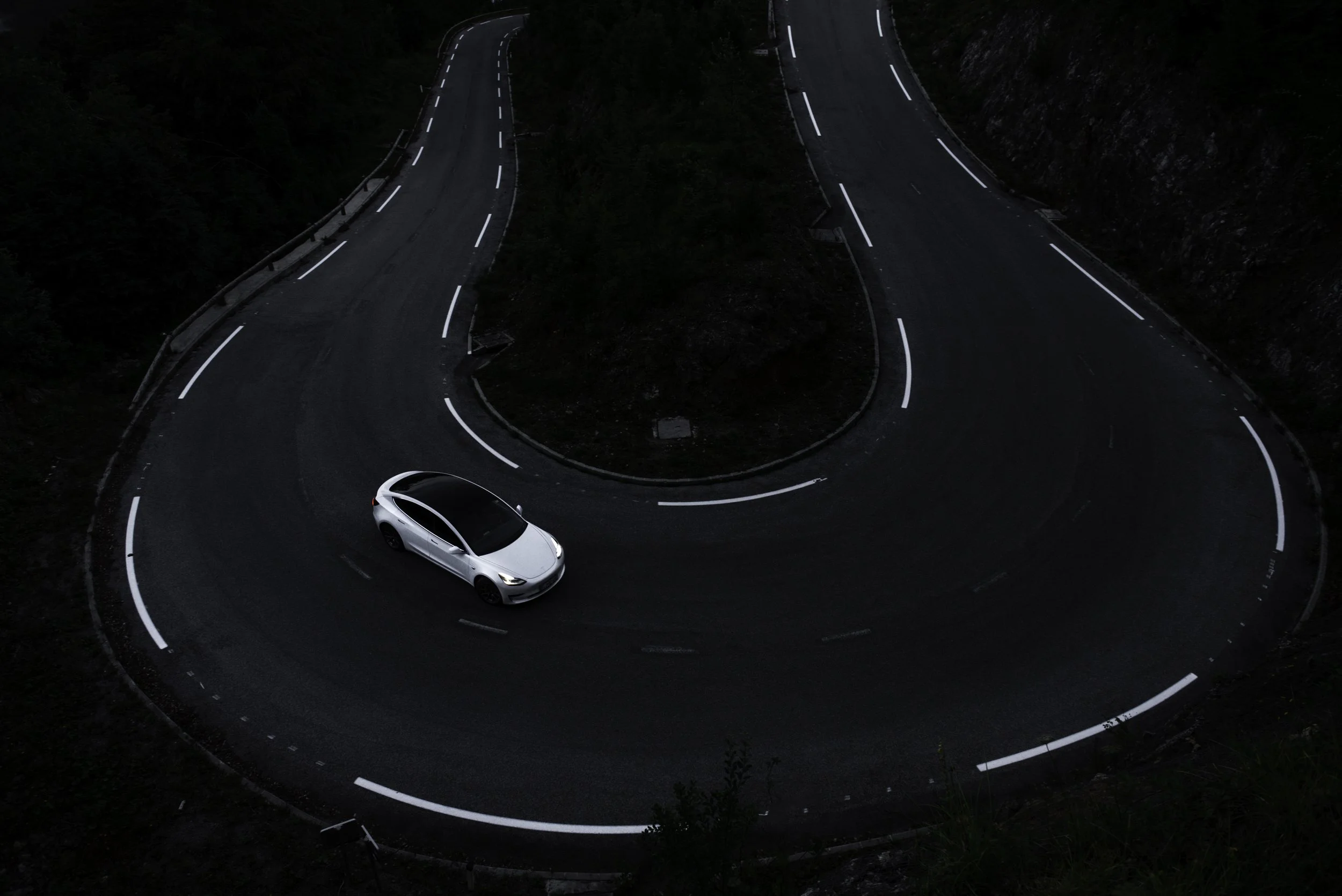 A white car driving on a winding mountain road at dusk or night, surrounded by dark terrain and steep cliffs.