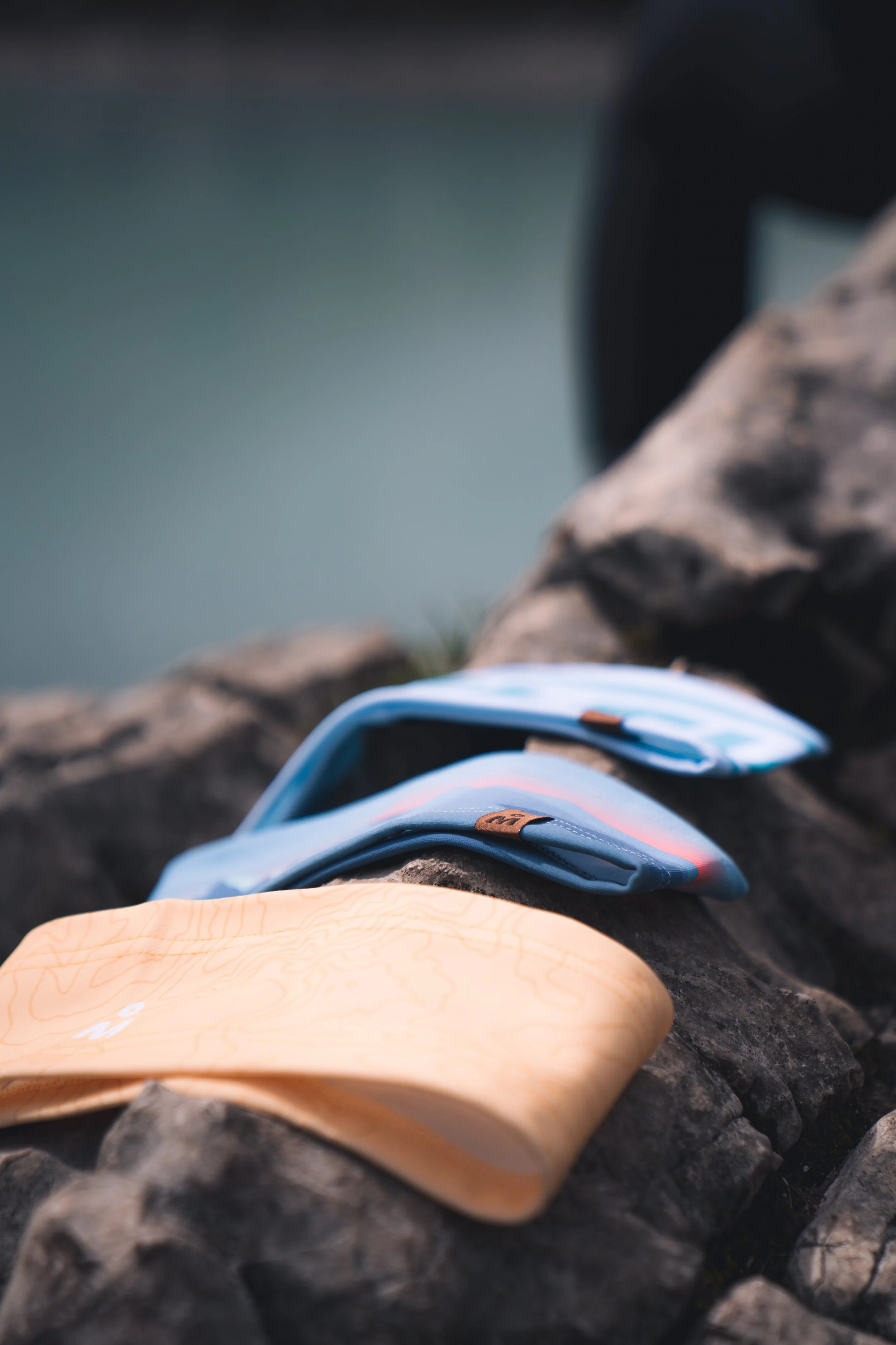 Climbing gear including a blue harness with brown accents and a beige climbing tape is laid out on dark rocks near a body of water.