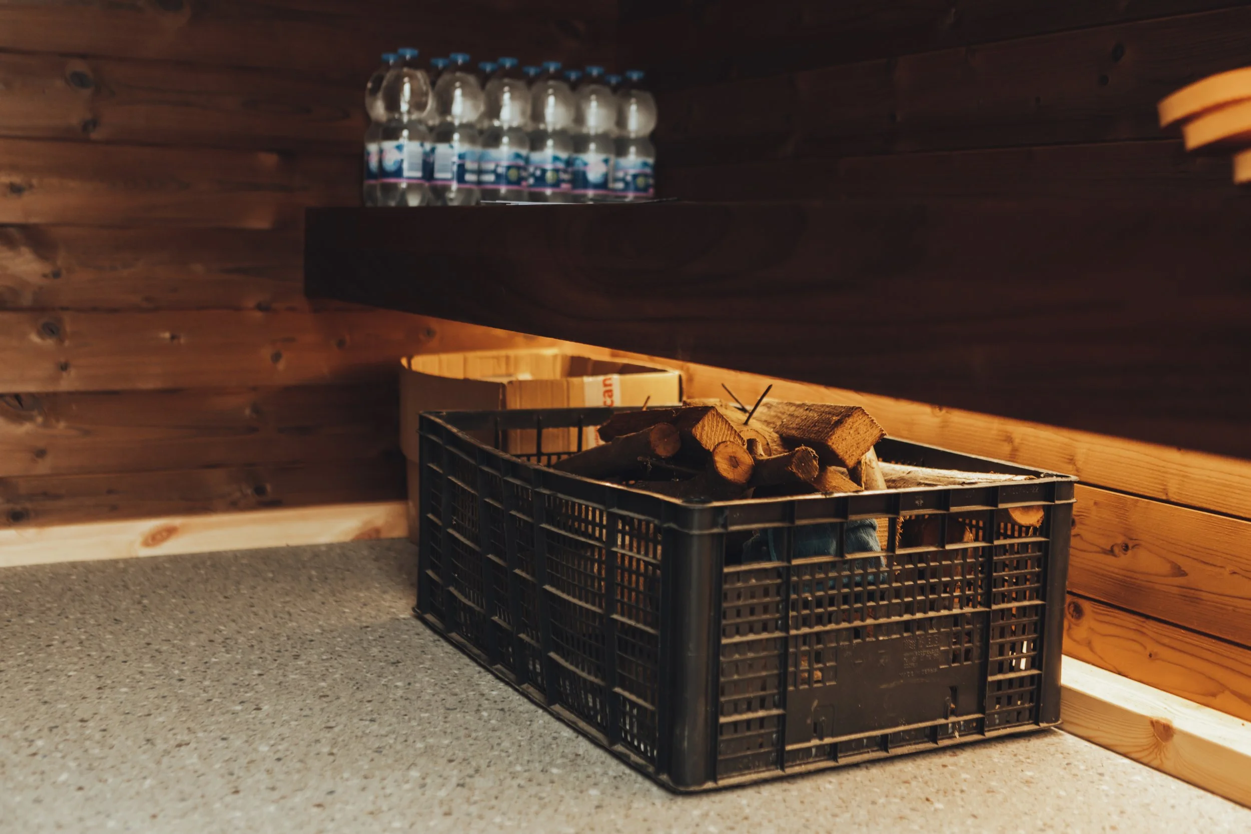 A black plastic crate filled with chopped firewood, placed on a grey carpeted floor against a wood-paneled wall inside a room.