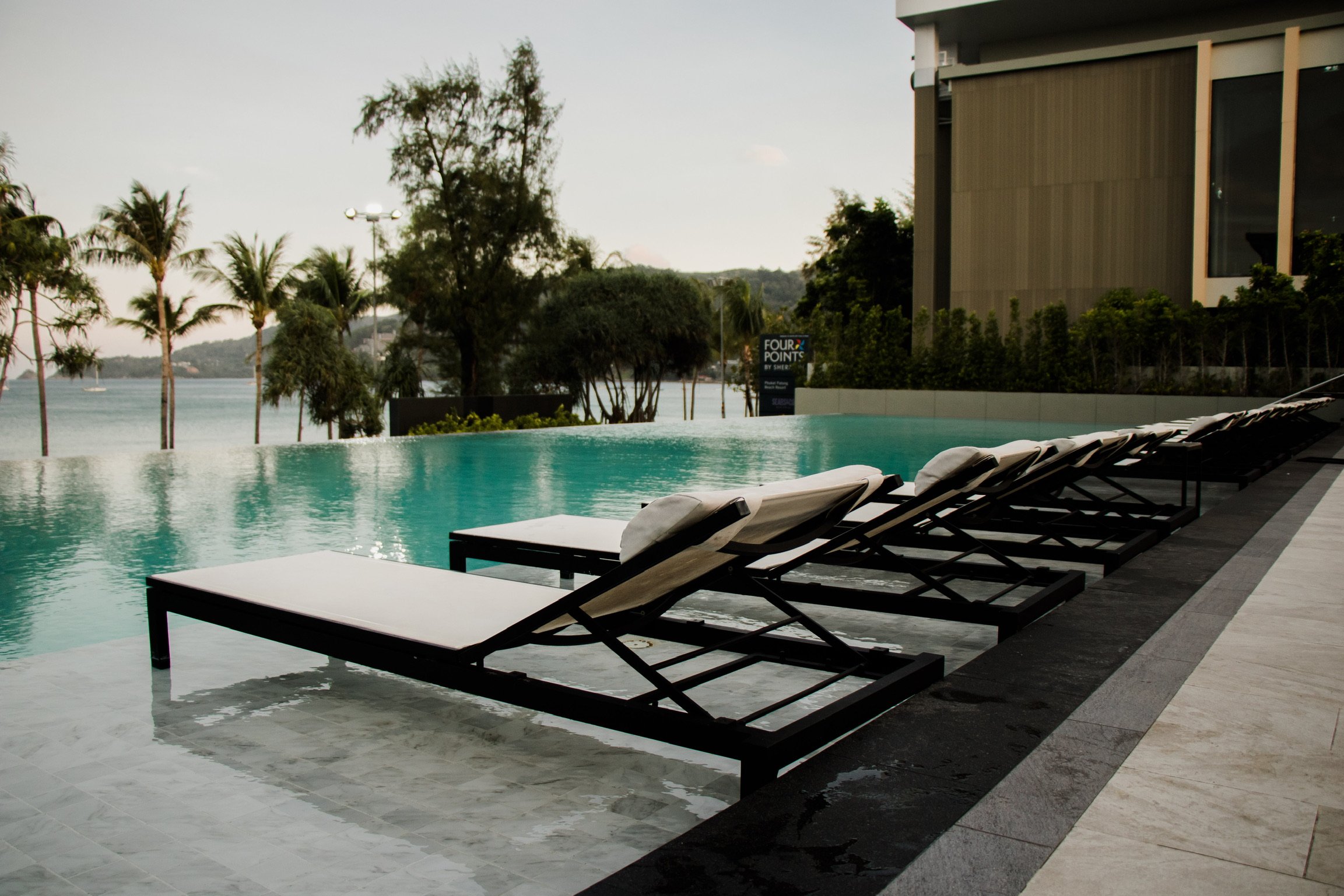 Six white lounge chairs with rolled towels on them are lined up next to an infinity pool with a view of trees and water in the background.