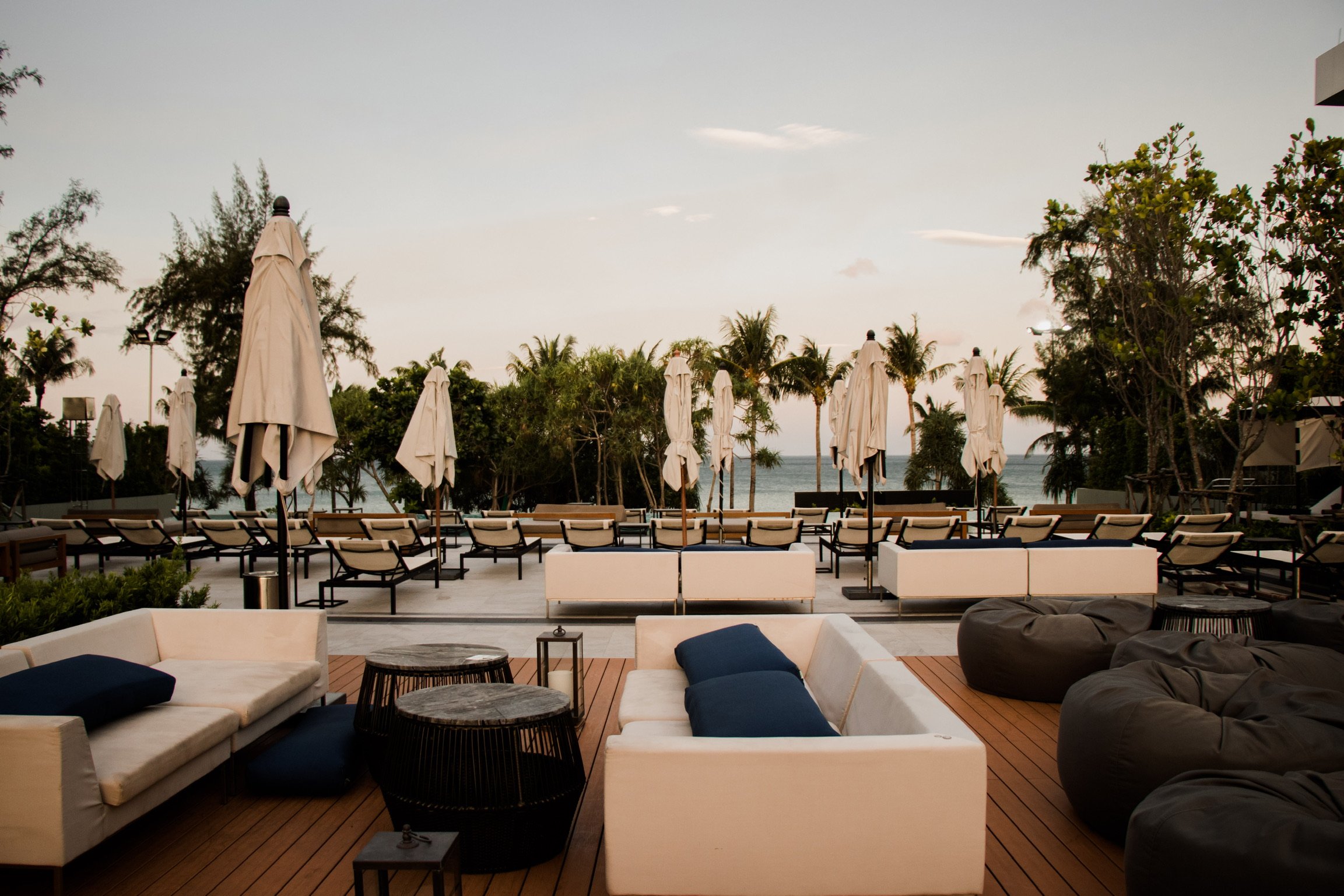 Empty poolside lounge area with white sofas, black bean bags, and closed umbrellas, overlooking the ocean with trees and palm trees in the background during early evening or late afternoon.