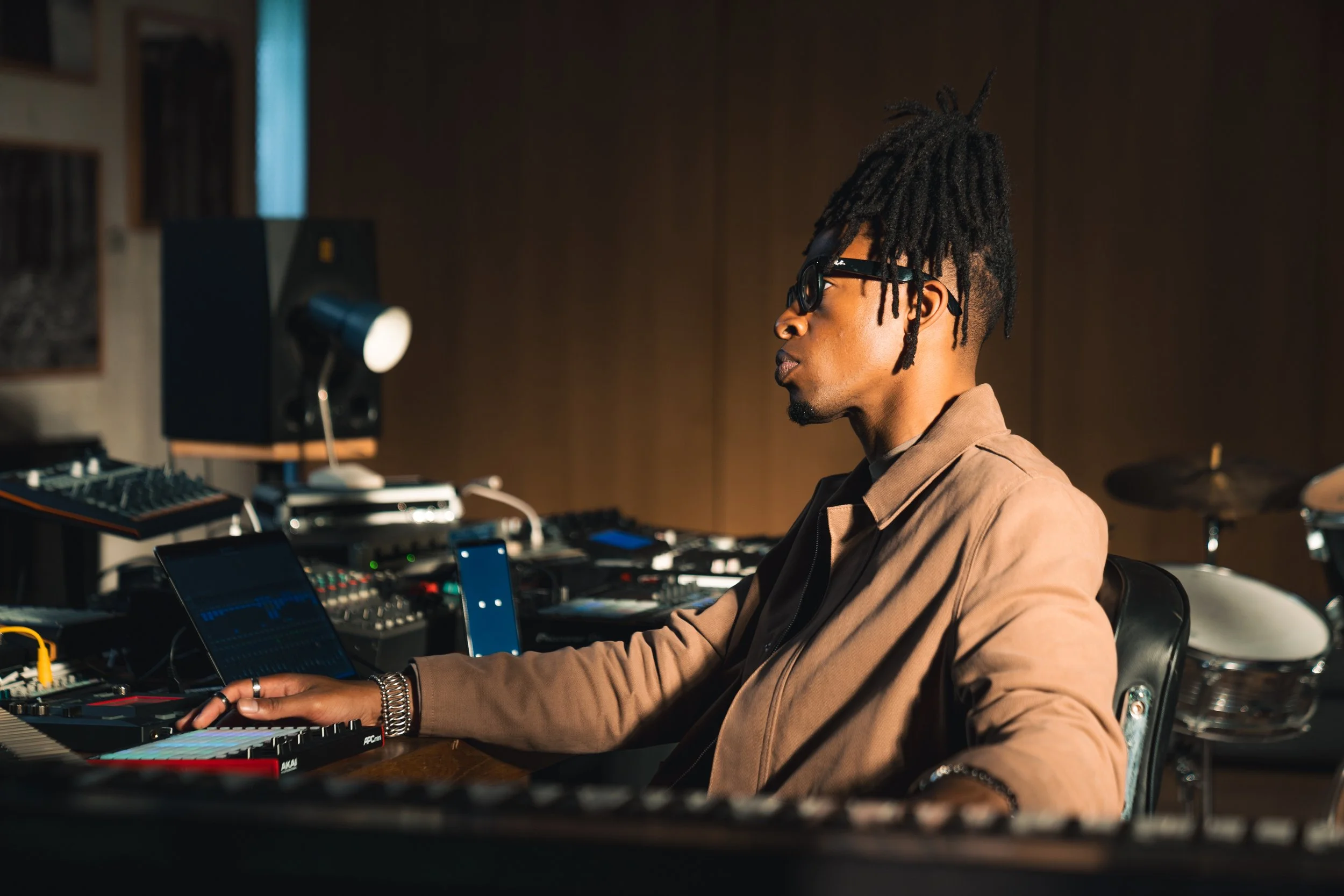 A man with dreadlocks, glasses, and dark skin working on music equipment in a studio, with various electronic music gear, keyboards, and drums around him.
