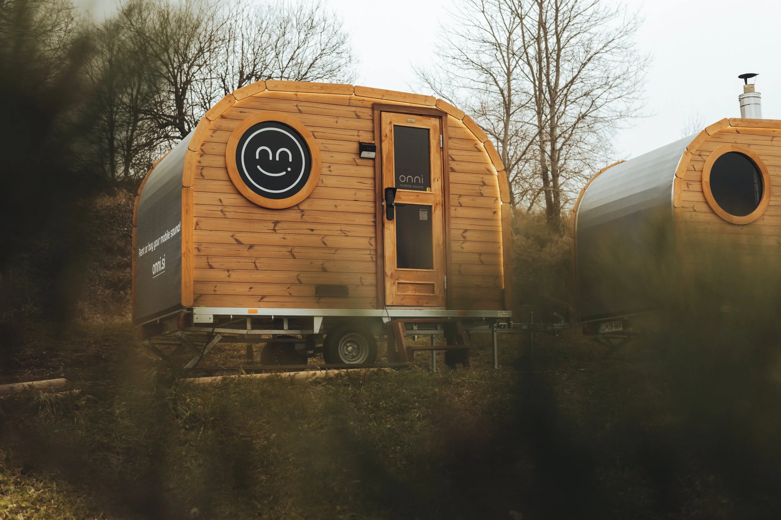 A small wooden mobile sauna trailer with a smiling face logo on the front and the text 'onni mobile sauna' on the door, parked outdoors with trees in the background.
