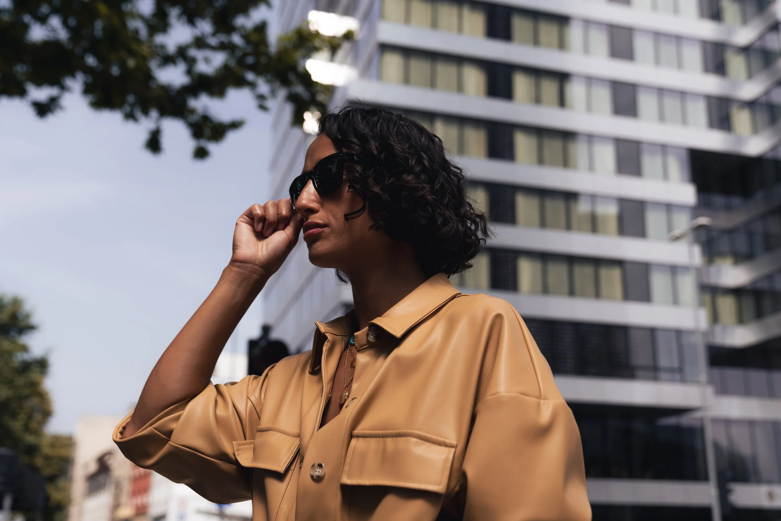 Woman with curly dark hair and sunglasses adjusting her glasses, wearing a beige leather jacket, standing outdoors in front of a modern high-rise building.