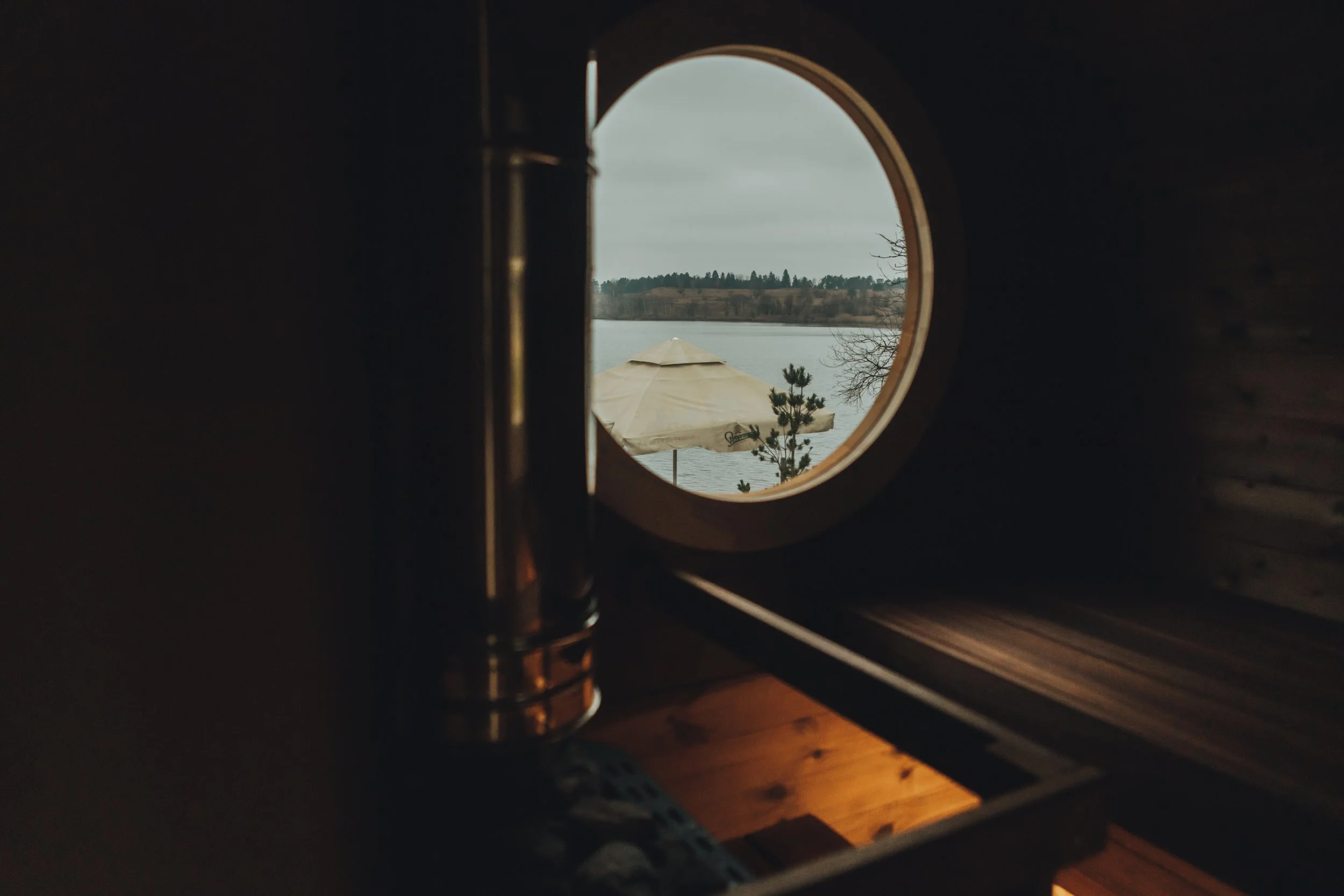 View through a round window showing an outdoor scene with a lake, a beige umbrella, some small trees, and a cloudy sky.