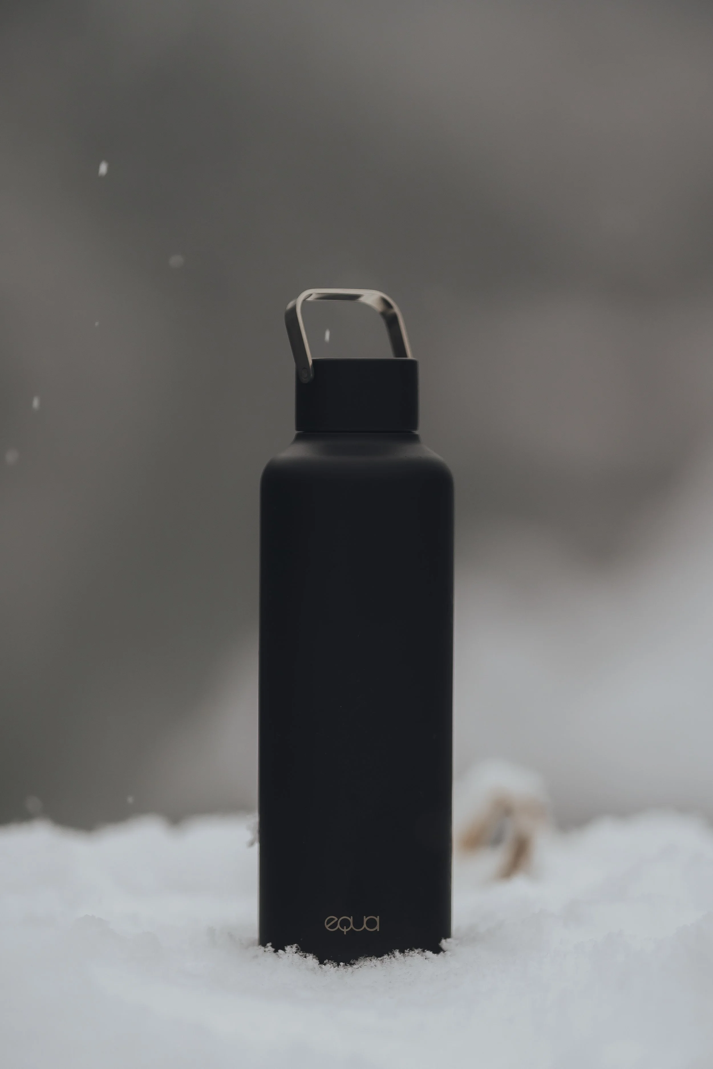 A black water bottle with a silver handle standing on snow.