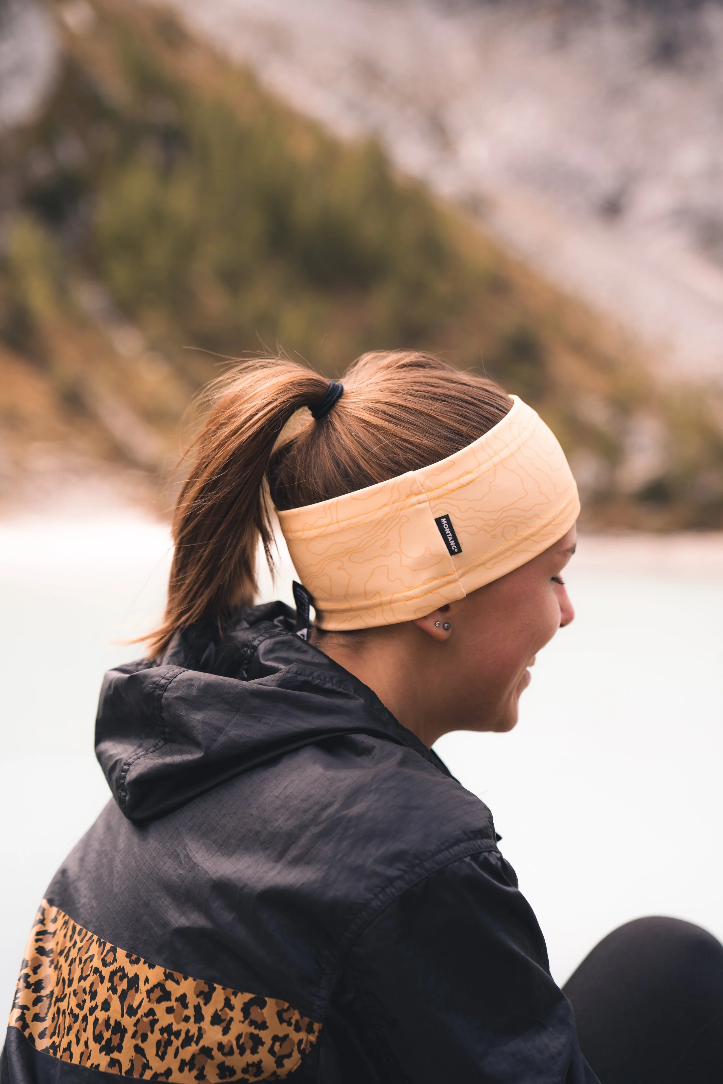 A woman with a ponytail wearing a yellow headband and a black jacket with a leopard print patch, sitting outdoors near water with blurred trees in the background.
