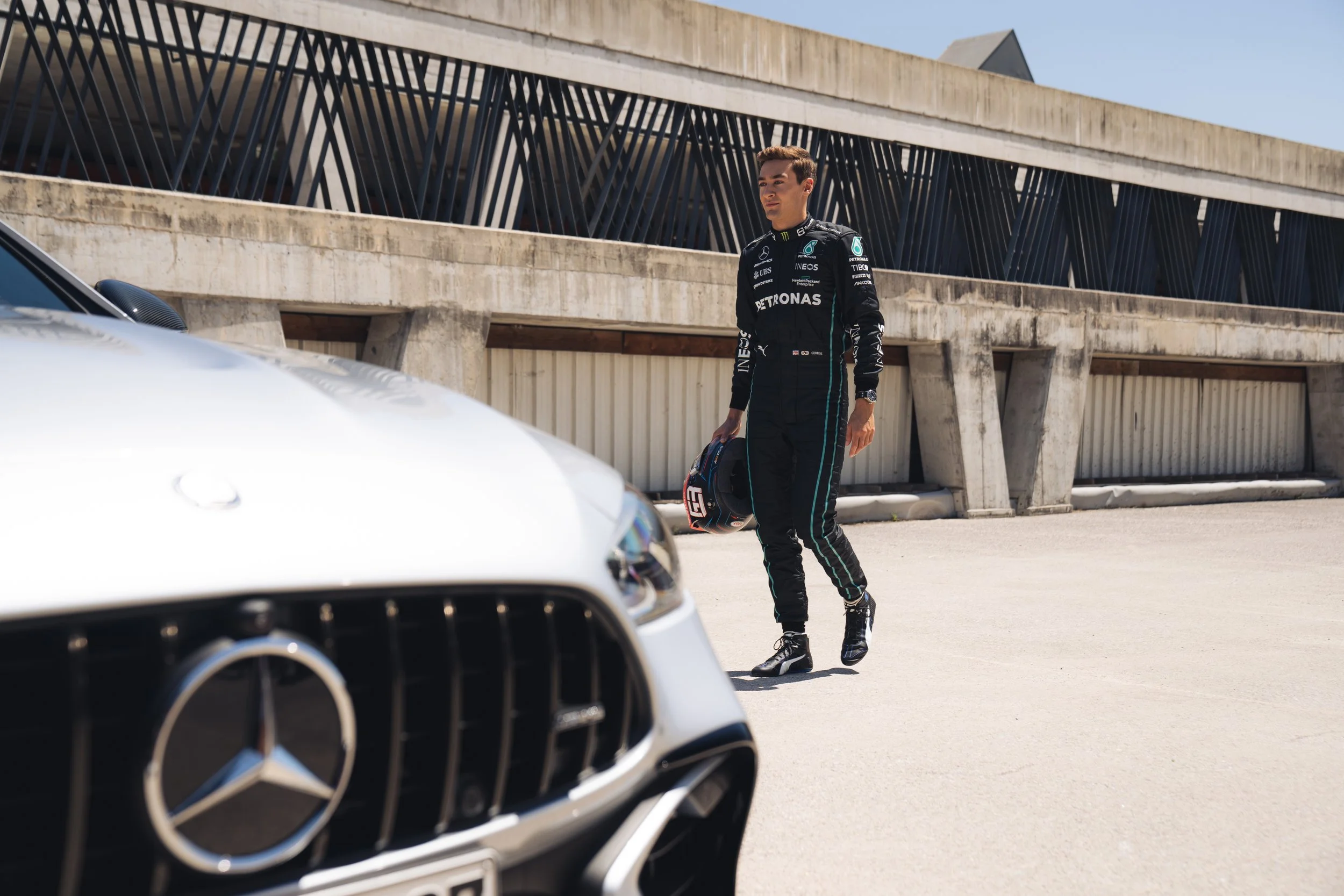 A race car driver dressed in a black racing suit holding a helmet, walking past a silver Mercedes-Benz sports car in a parking lot near a modern building.