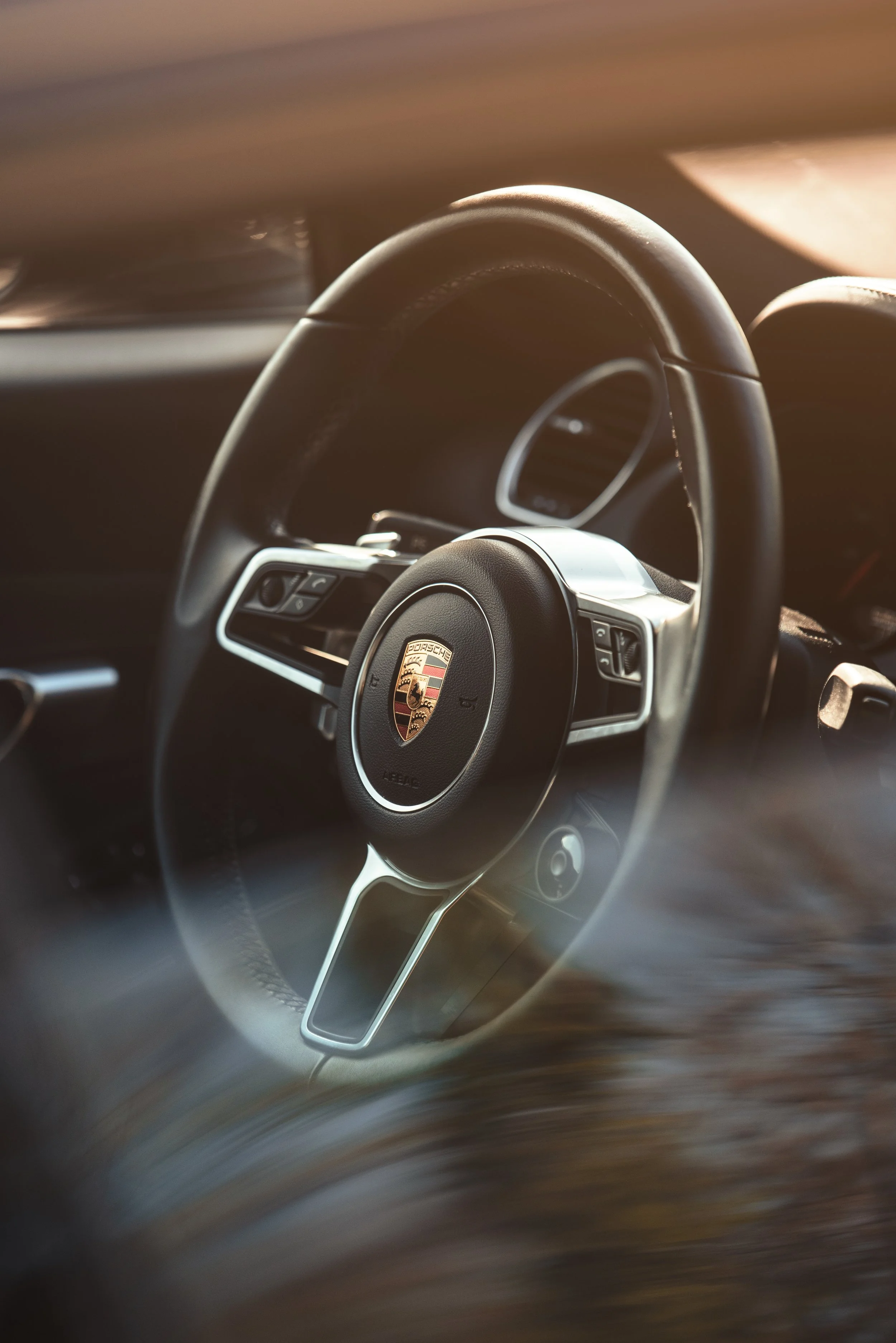 Close-up of a Porsche car steering wheel with the Porsche logo in the center, interior of the car visible with blurred background.