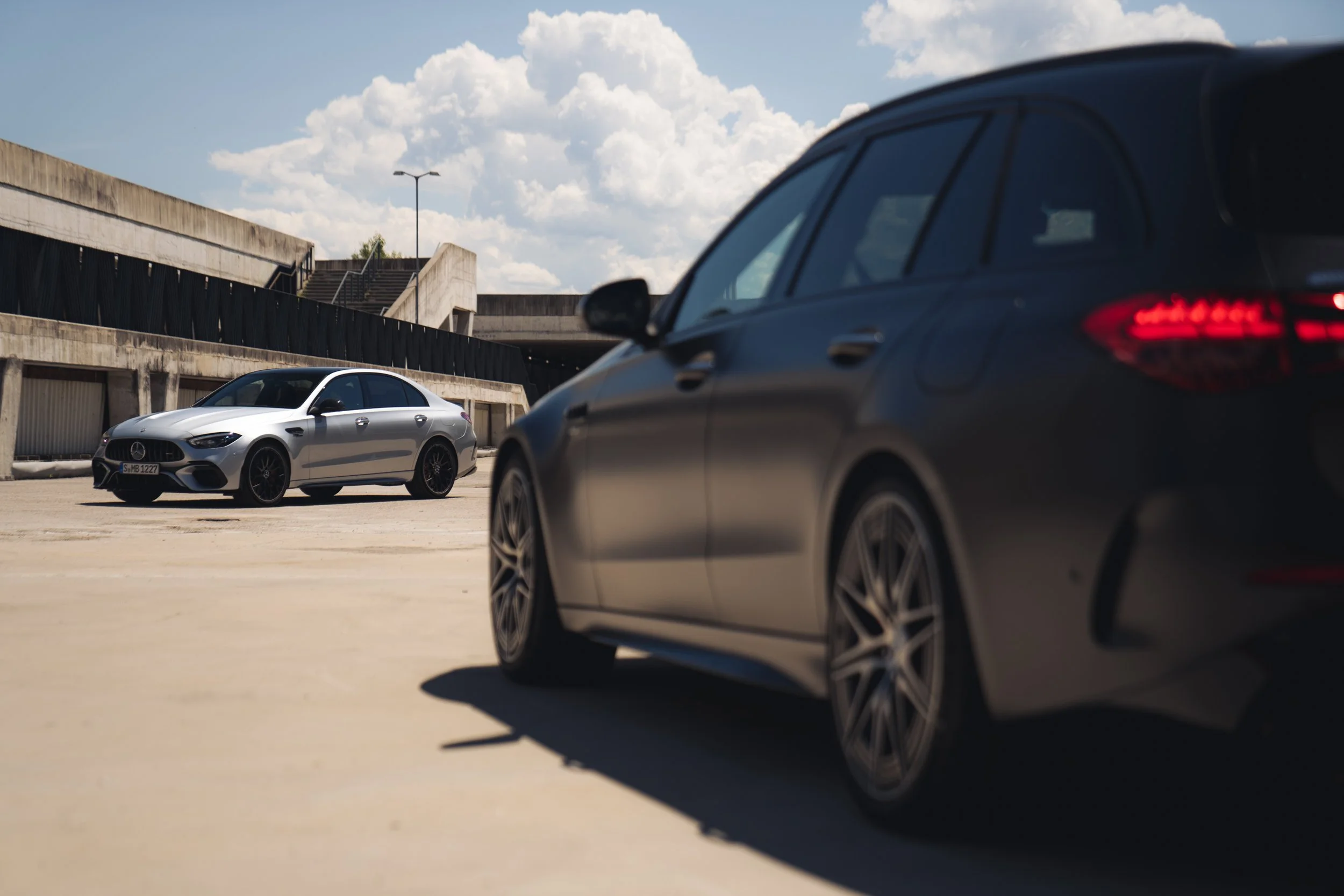 Two luxury cars, one black and one silver, parked in an open lot with a modern concrete building and stairs in the background under a partly cloudy sky.