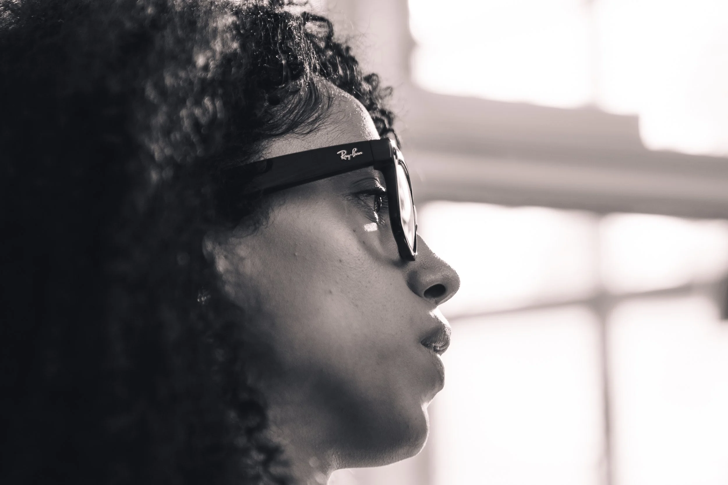 Close-up side profile of a woman wearing Ray-Ban sunglasses, with curly hair, looking contemplative in black and white.