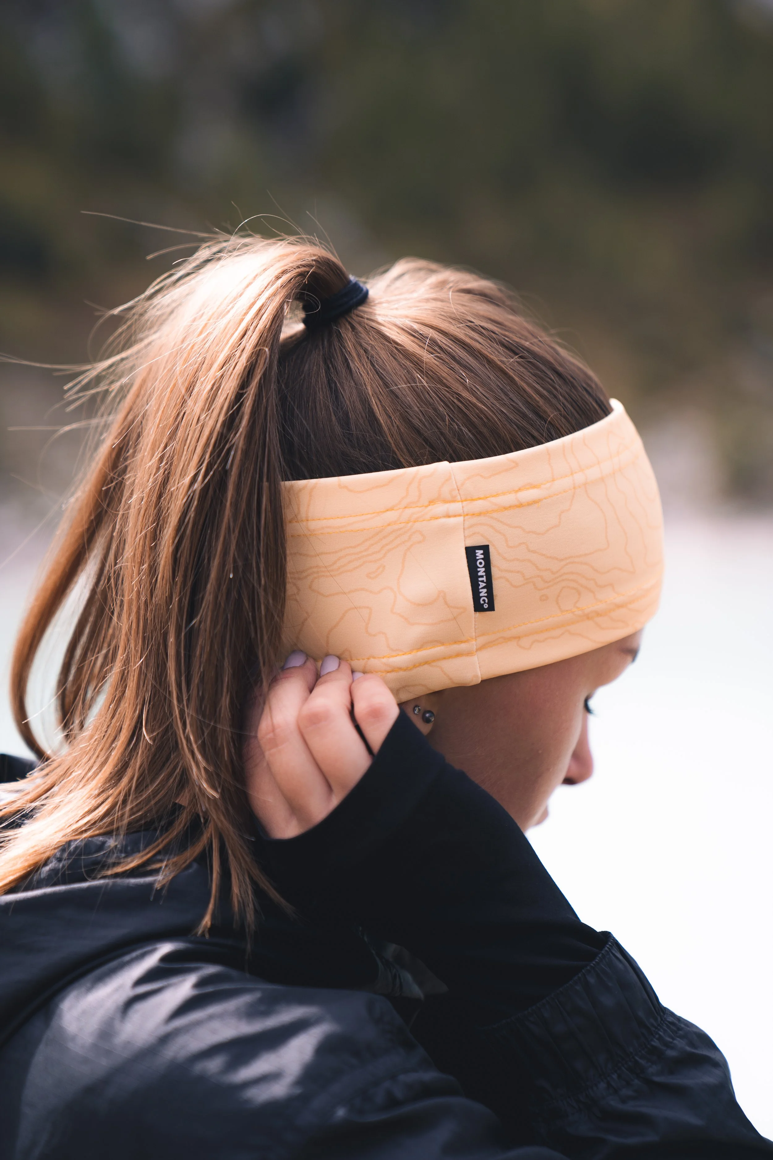 A woman outdoors wearing a yellow headband and adjusting an ear warmer, with brown hair tied back and a black jacket.