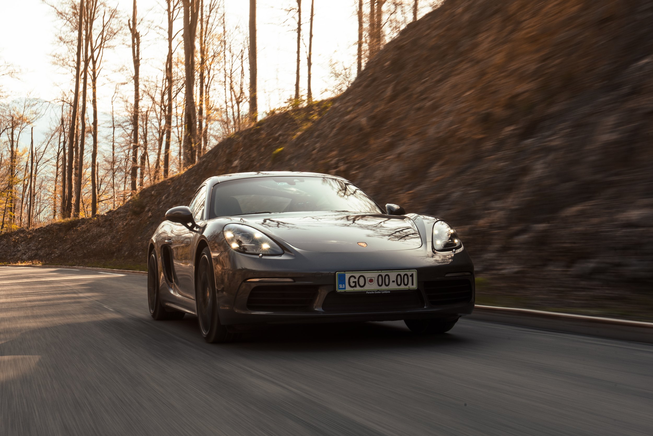 Black Porsche sports car driving on a winding mountain road with trees and rocky hillside in the background.