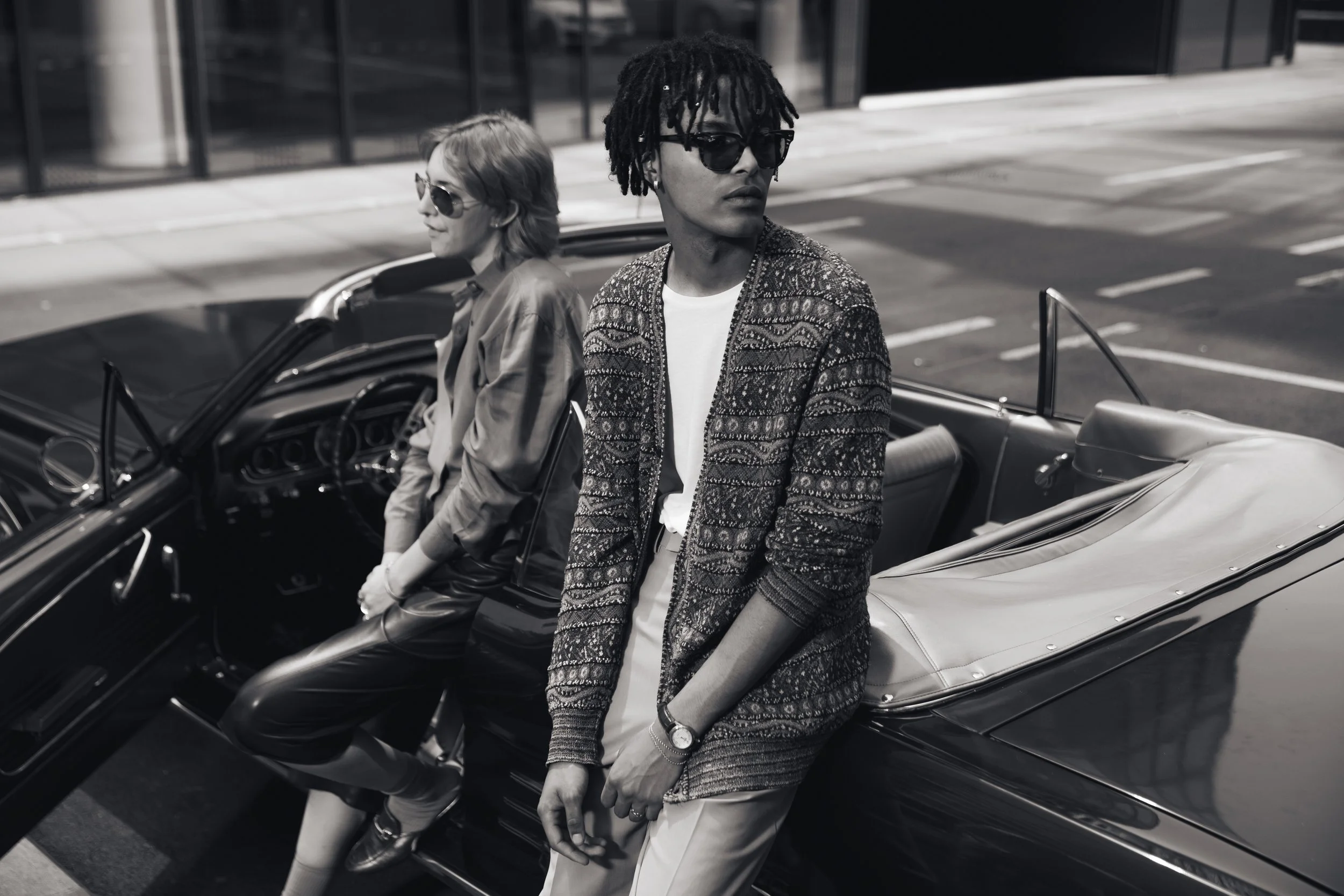 Two stylish individuals with sunglasses are near a vintage convertible car on a city street, one leaning against the car and the other sitting inside.