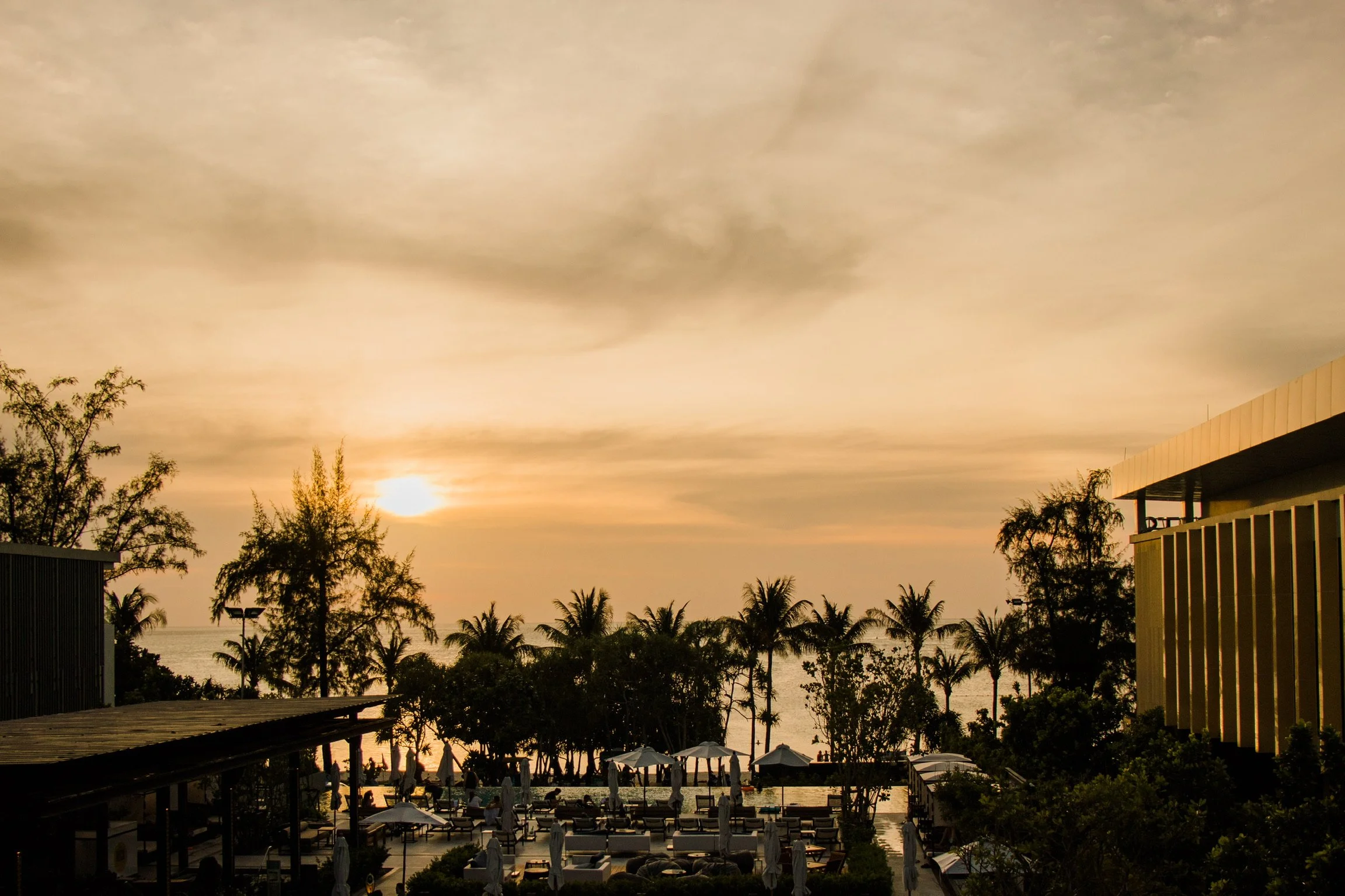 Sunset view over a tropical resort with palm trees, outdoor seating, umbrellas, and a view of the ocean in the background.