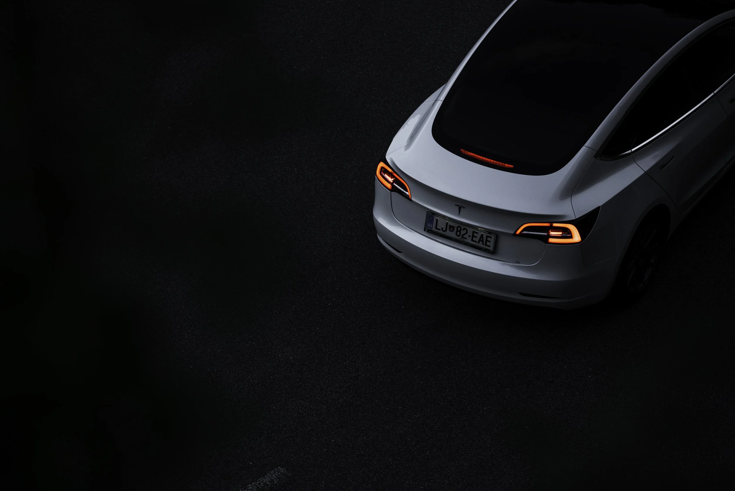 A white Tesla Model S electric car parked on a dark road at night, viewed from above. The vehicle's taillights are illuminated, and the license plate reads LJ 82 EAE.