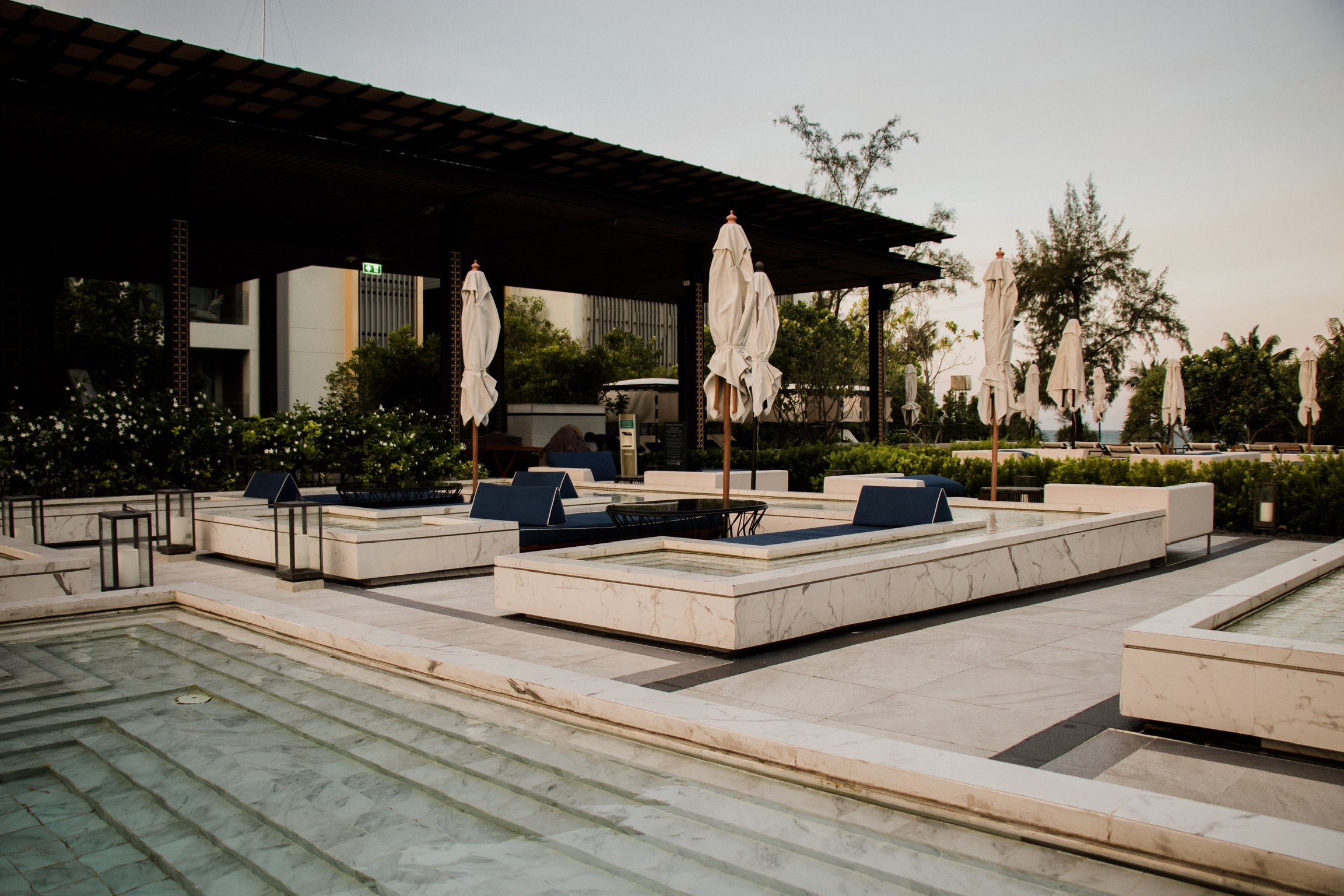 Empty outdoor lounge area with marble seating, folded umbrellas, and a covered shaded structure, surrounded by greenery and trees.