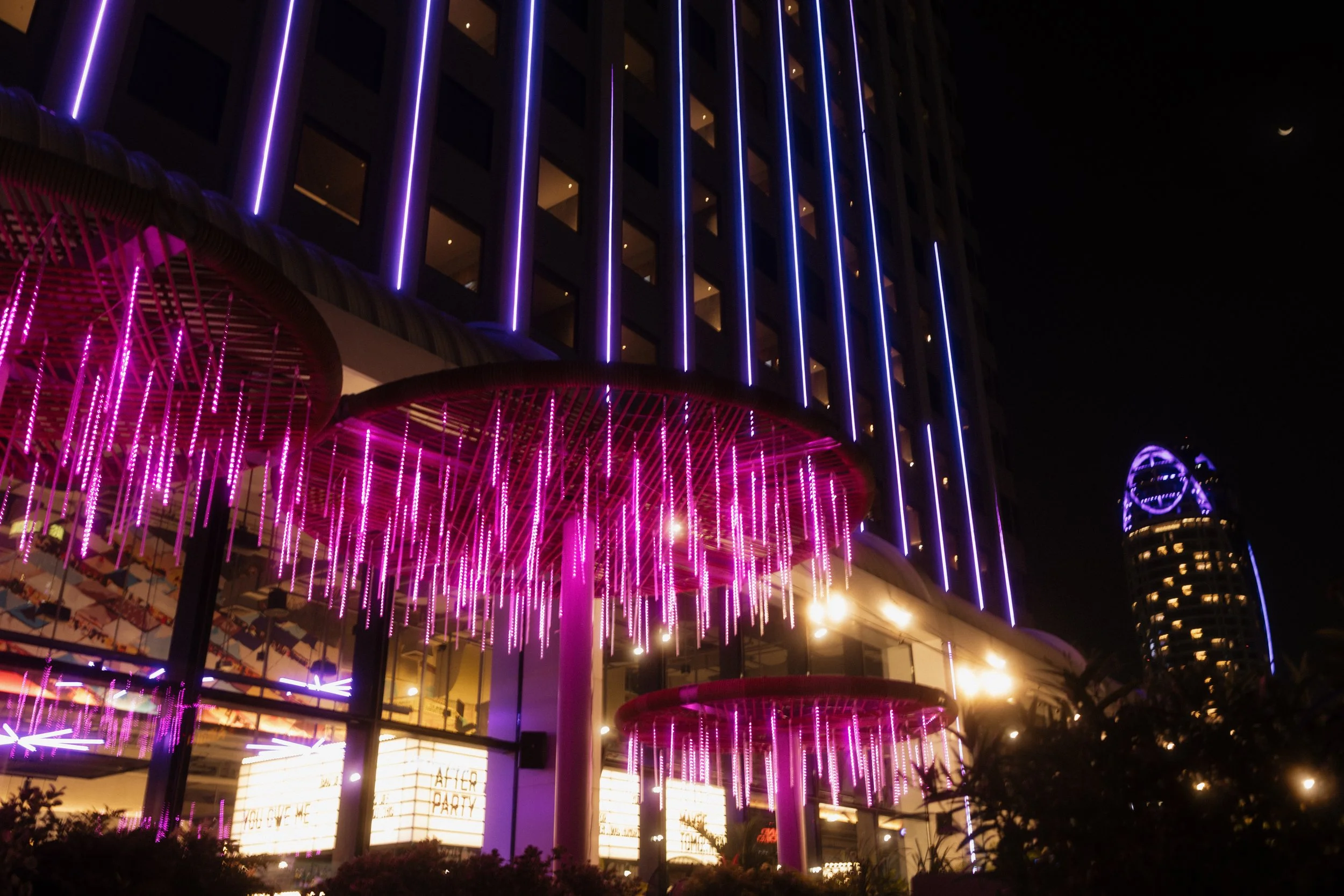 Night view of a modern building decorated with pink and purple neon lights, with a sign reading 'After Party' visible at the entrance.