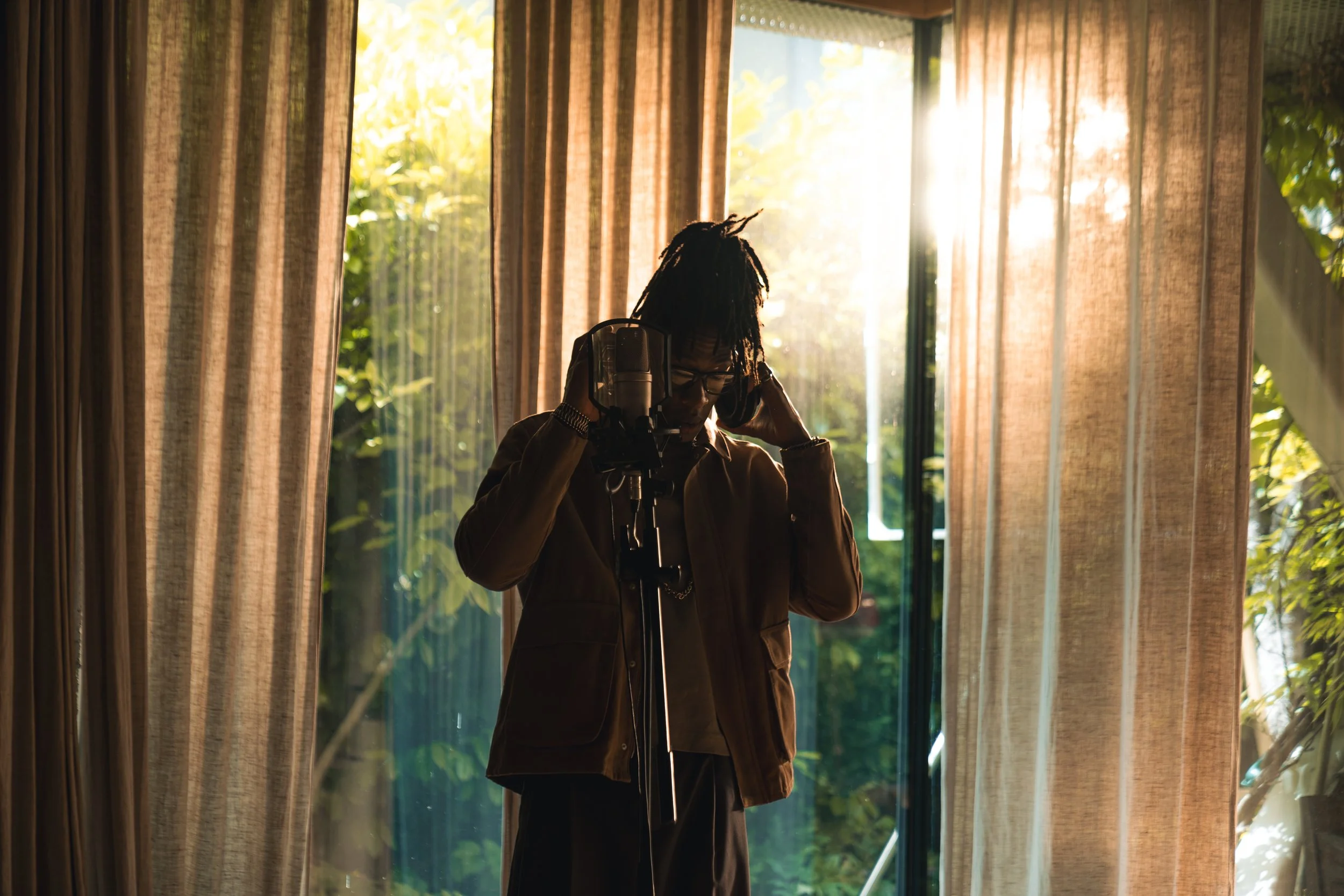 Person with dreadlocks wearing glasses speaking into a microphone in a room with large windows and curtains, illuminated by sunlight and surrounded by outdoor greenery.