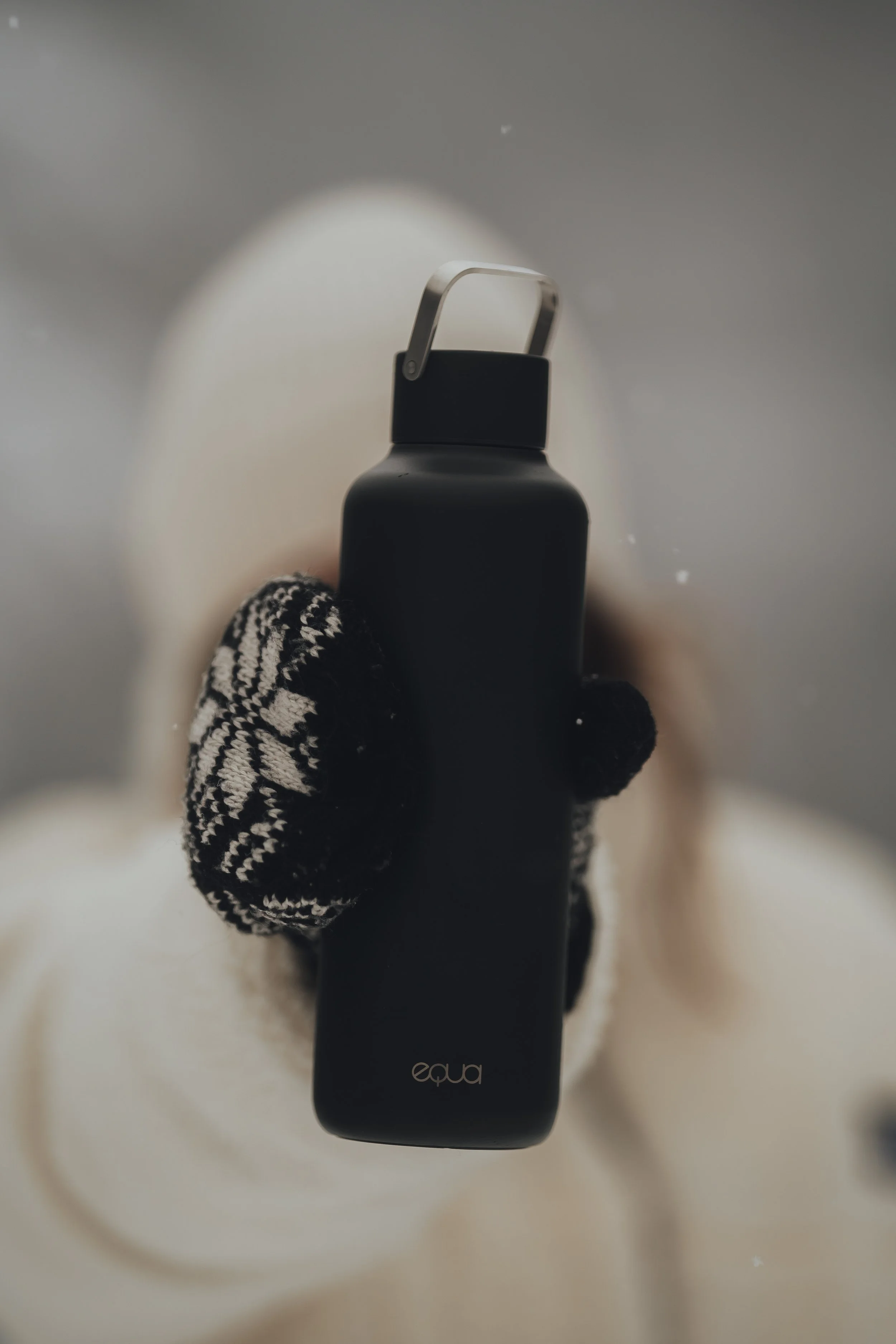 A person wearing black and white patterned gloves holding a black water bottle in front of their face.