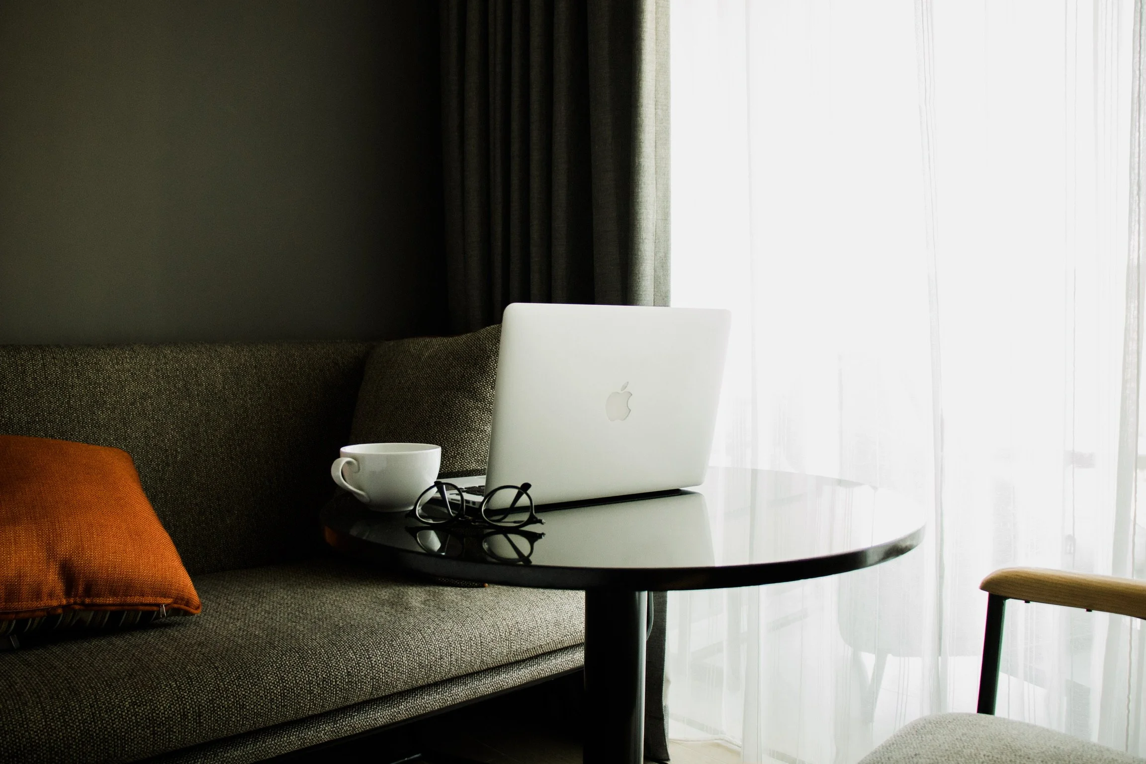 A living room scene with a sofa, an orange pillow, a black round coffee table, a silver laptop, a white cup, a pair of glasses, and a wooden chair with a light-colored cushion. There are dark curtains and a bright window with sheer white curtains in 