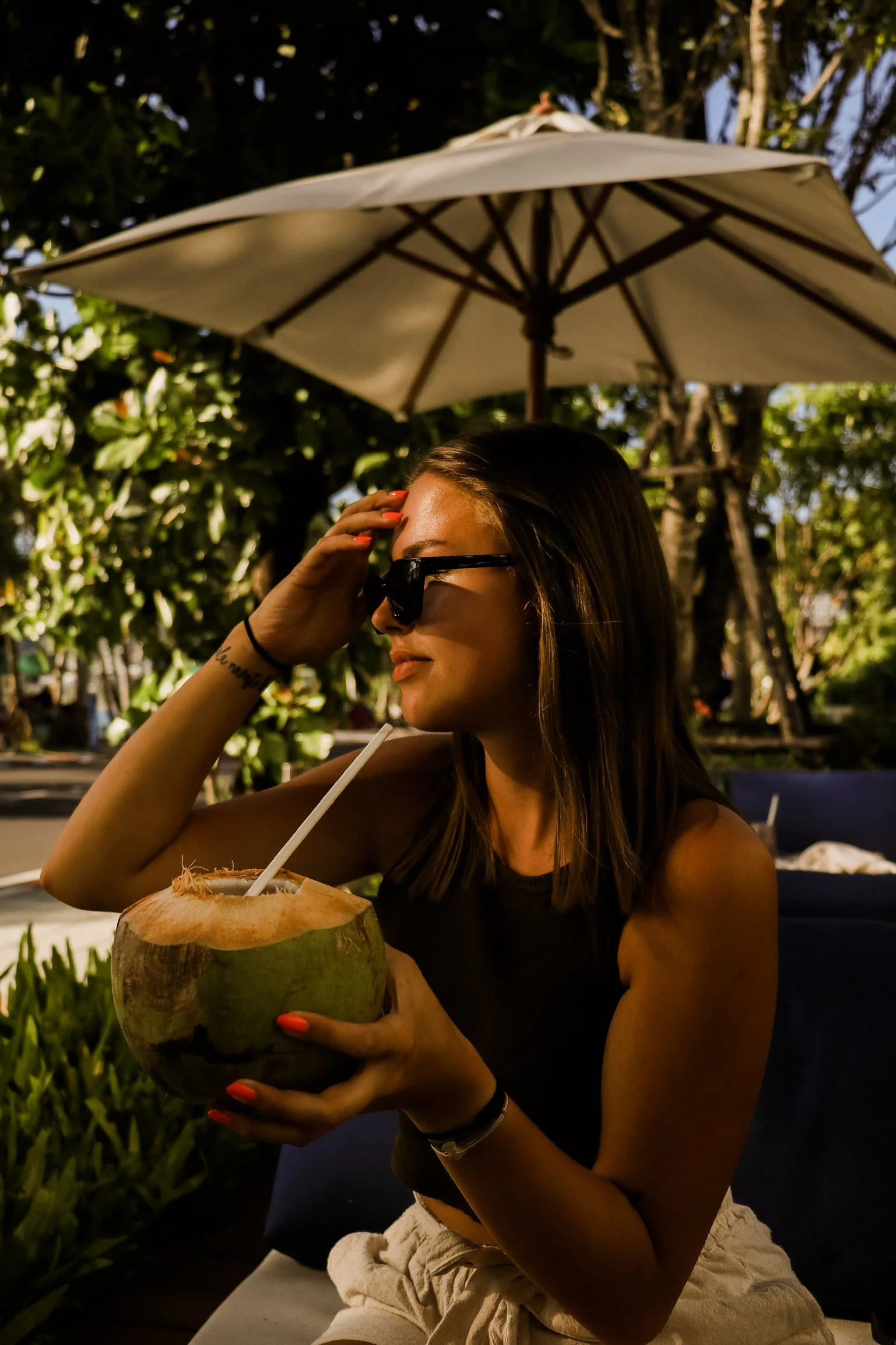 Woman sitting outdoors holding a coconut with a straw and wearing sunglasses, under a large umbrella.