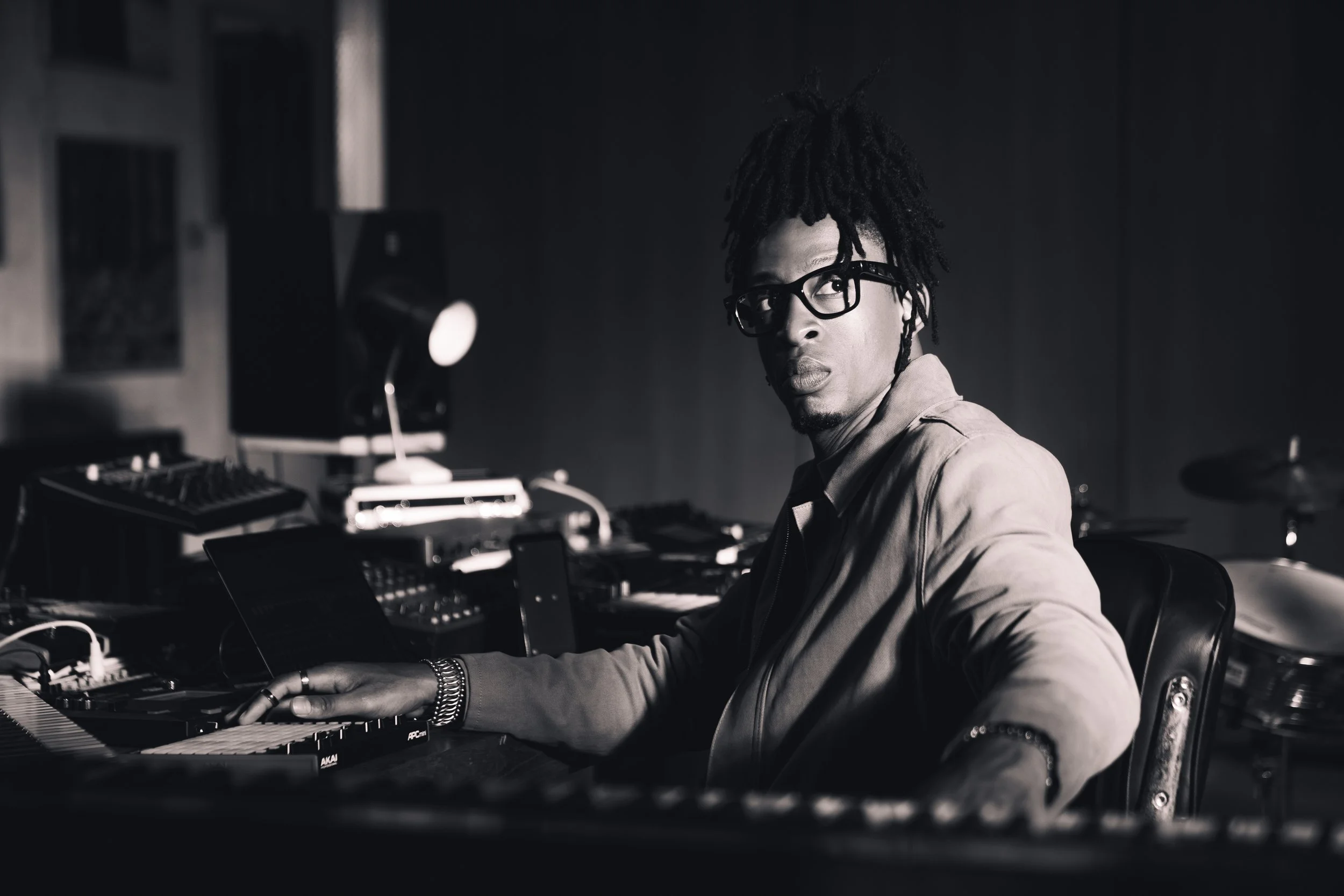 A man with dreadlocks and glasses sitting at a keyboard, recording studio with music equipment in the background, black and white photo.