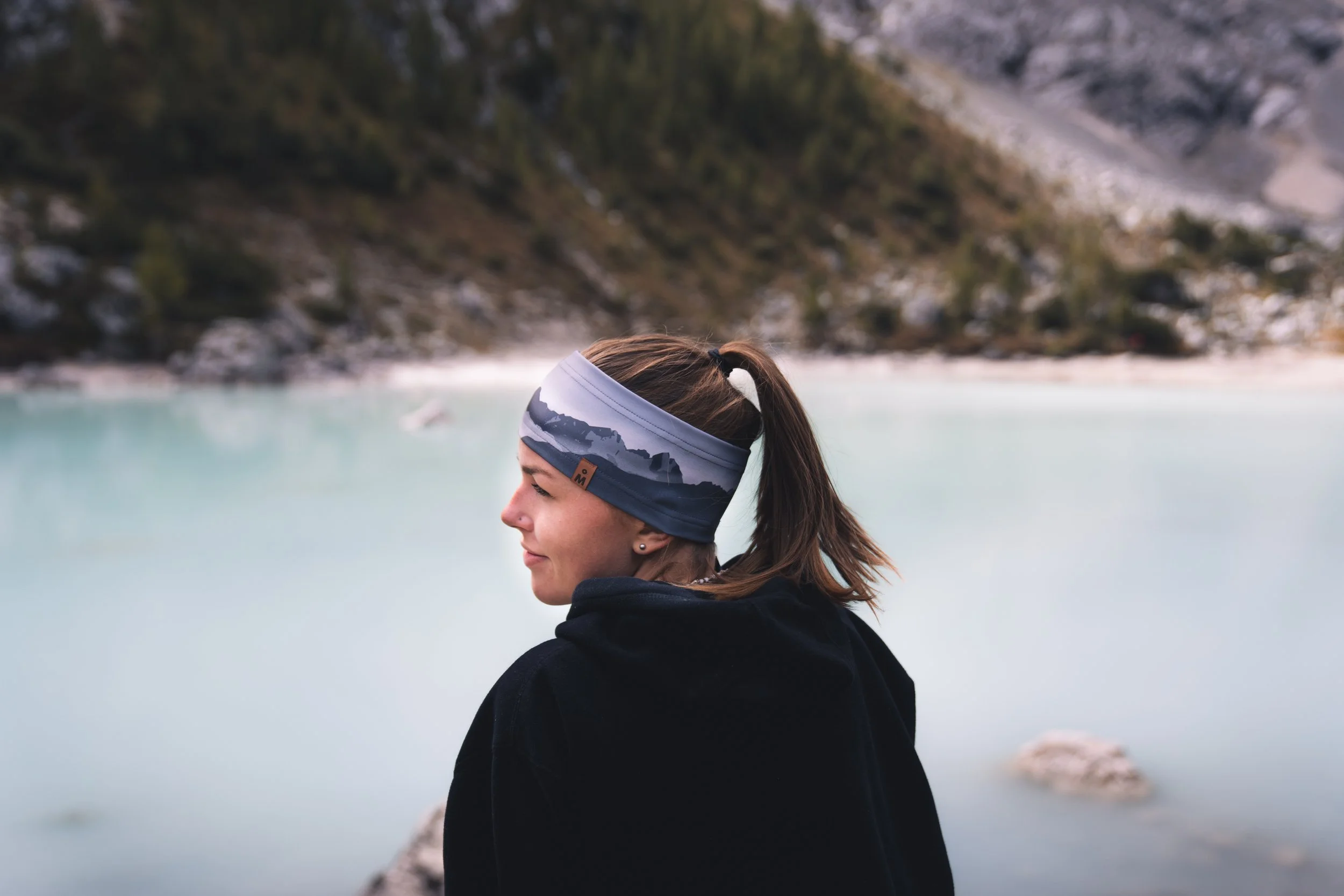 Woman with a headband by a mountain lake, facing left, wearing a black hoodie.