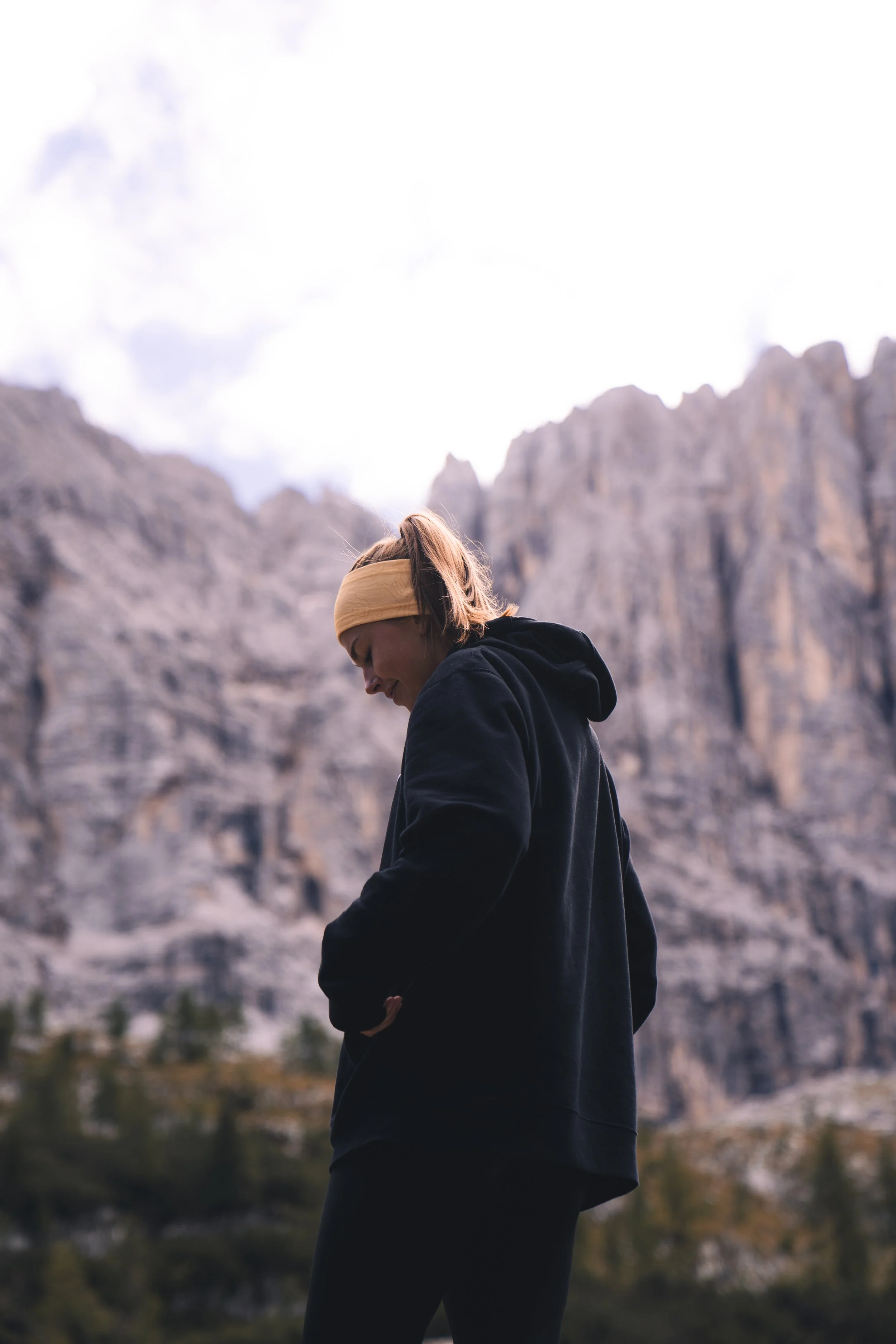 A person wearing a black hoodie and a tan headband standing outdoors in front of a mountain range with a cloudy sky.