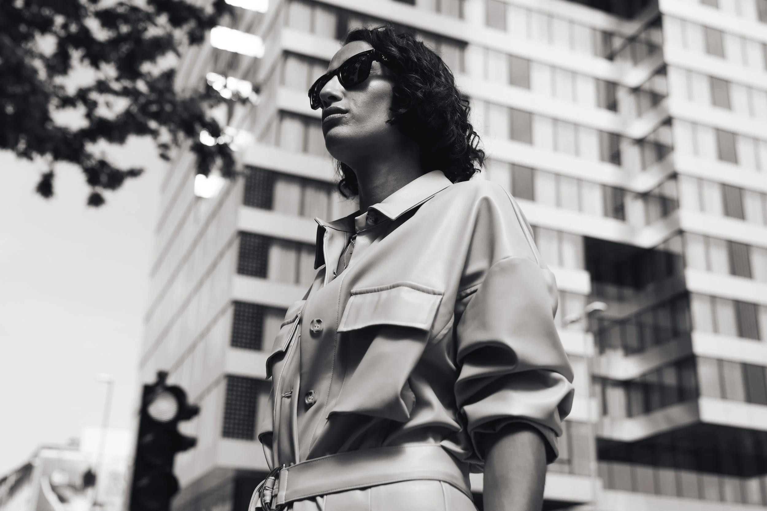 A woman wearing sunglasses and a leather jacket standing outdoors in front of a modern, multi-story building.