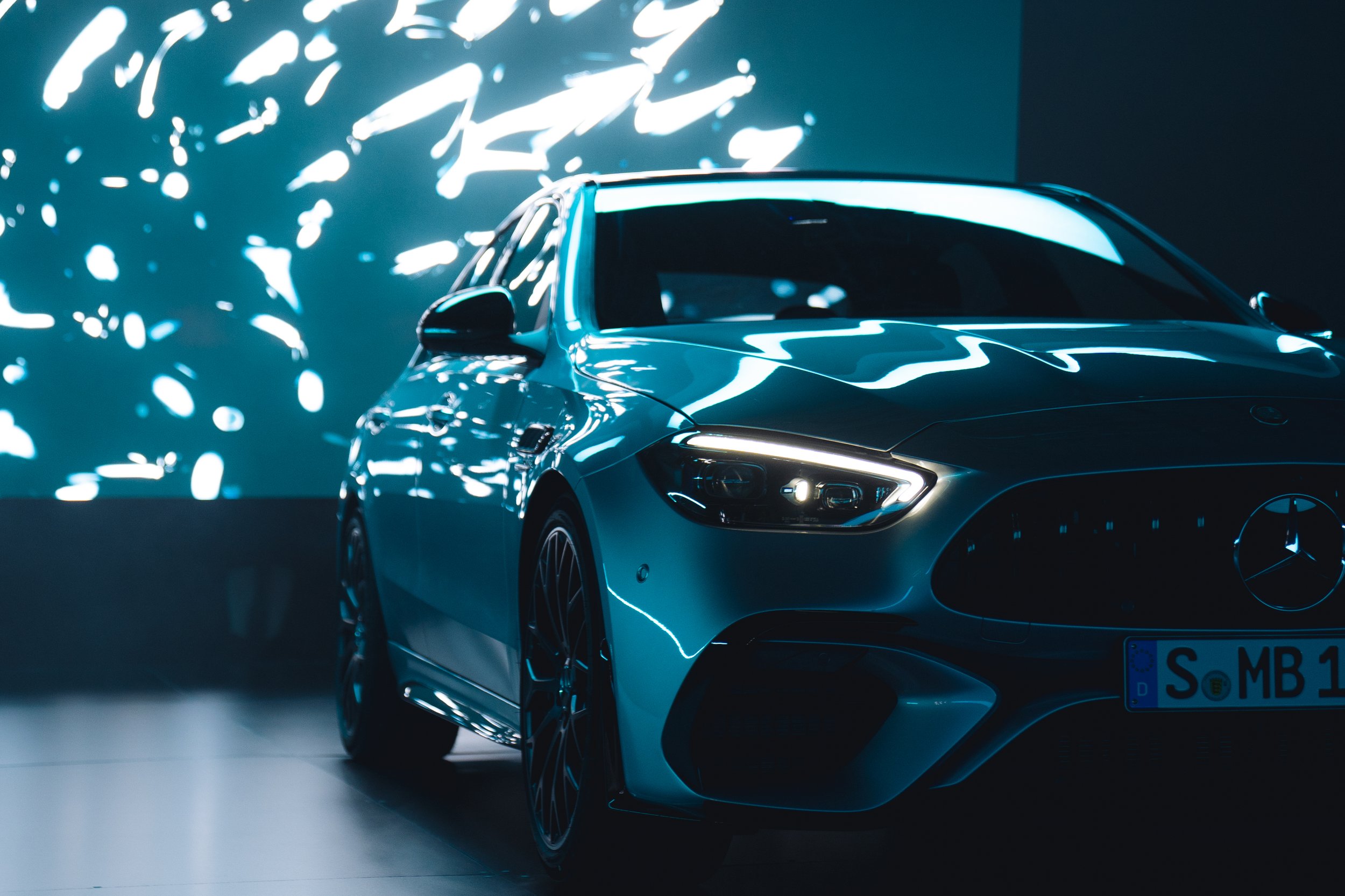A sleek blue Mercedes-Benz car displayed in a modern showroom with abstract light reflections on the wall behind it.