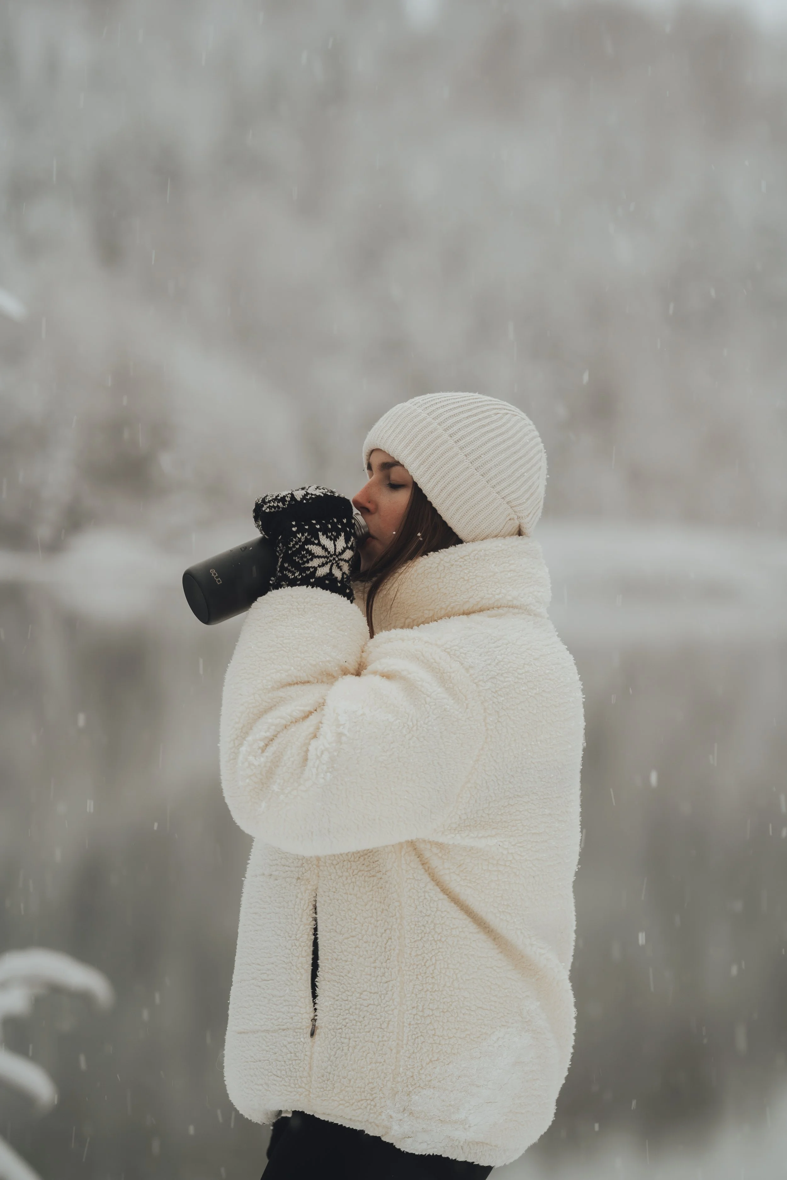 Woman in winter clothing drinking from a thermos outdoors in snowy weather.