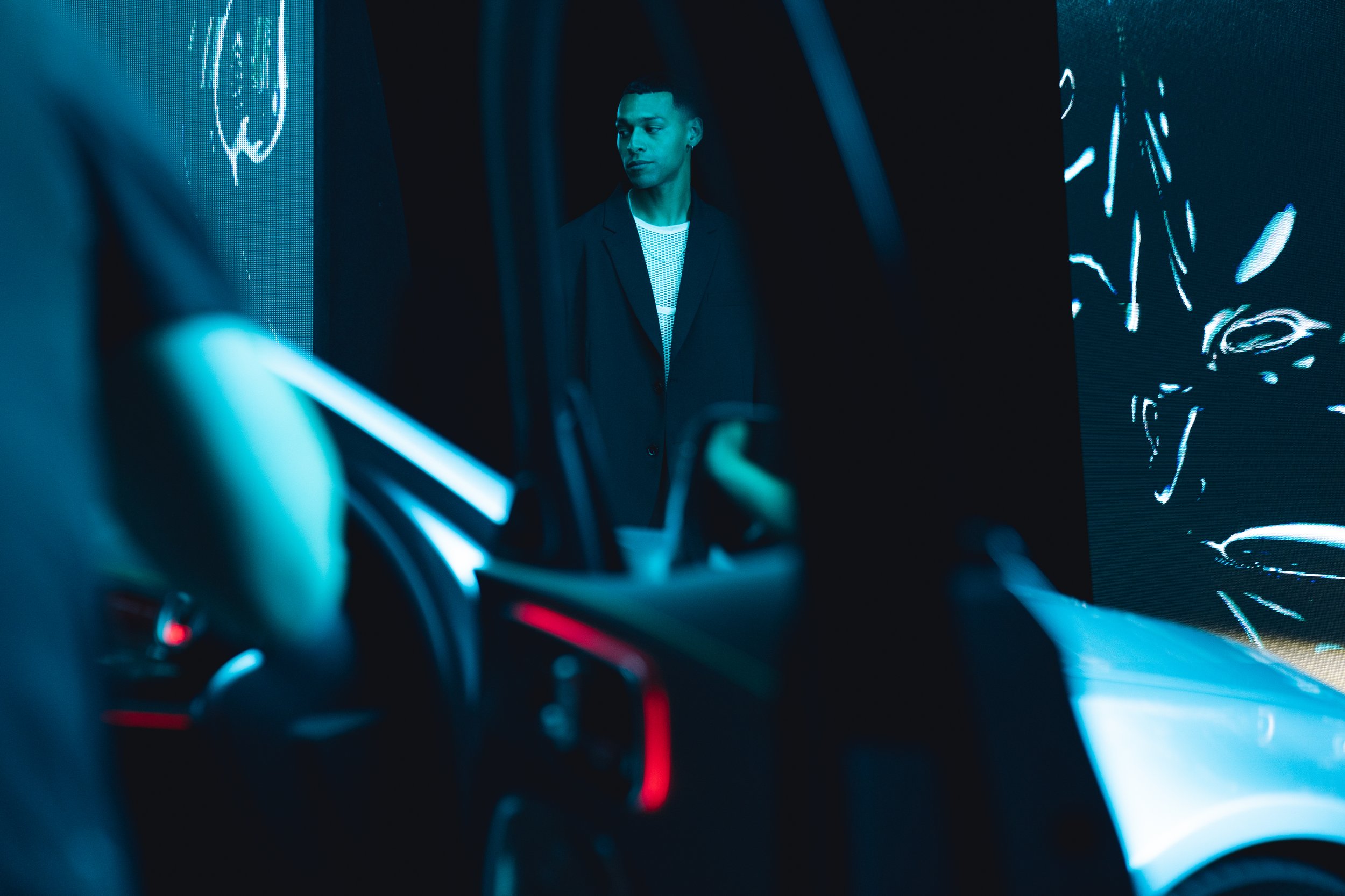 A man in a black blazer standing in a dark, futuristic environment with digital screens and neon lighting.