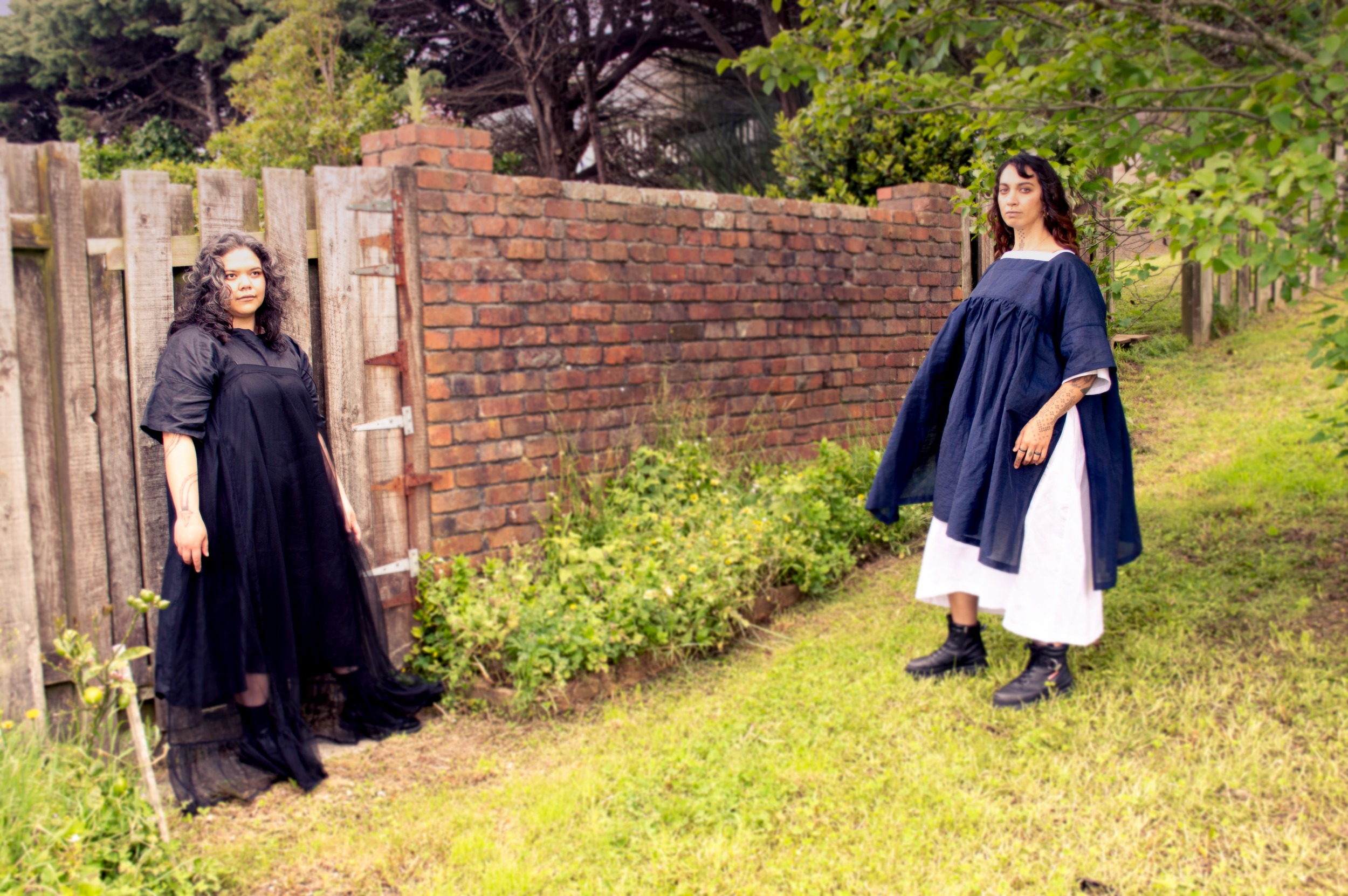 Two women standing outdoors in a garden. One woman is on the left near a wooden fence, wearing a black dress, with curly hair. The other woman is on the right near a brick wall, wearing a white dress with a blue overlay, and has tattooed arms, with dark hair.