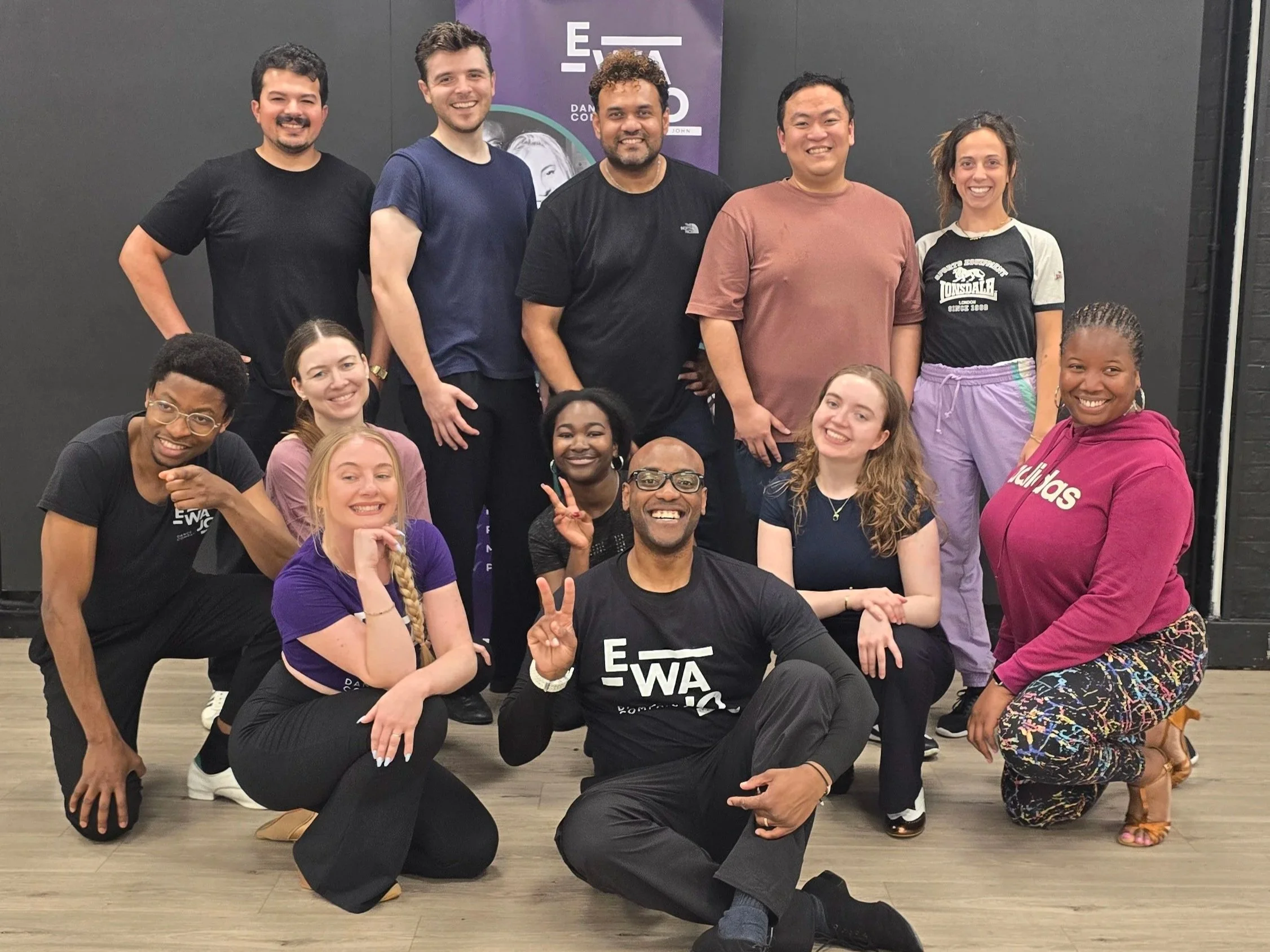 A group of people smiling and posing for a photo in a salsa dance studio with a black wall and purple banner in the background.