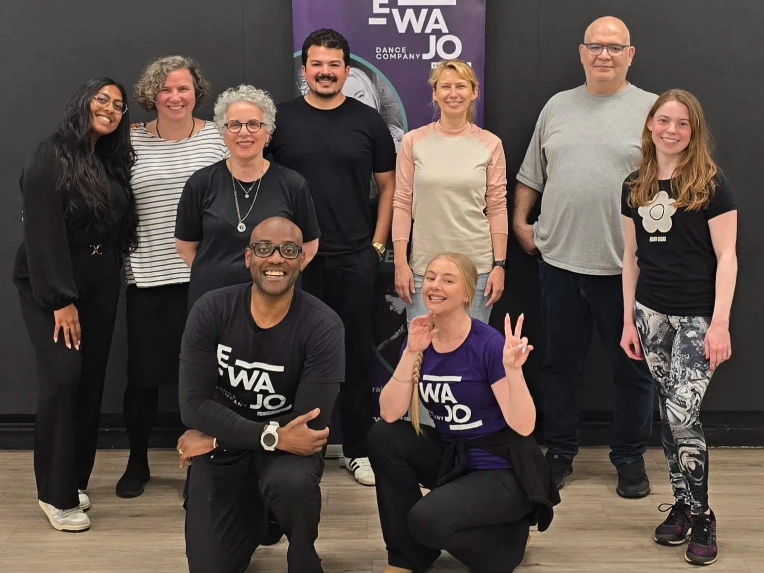 A group of ten people posing together indoors, with some wearing salsa dance company shirts and a dance company banner in the background. They are smiling and making various hand gestures.