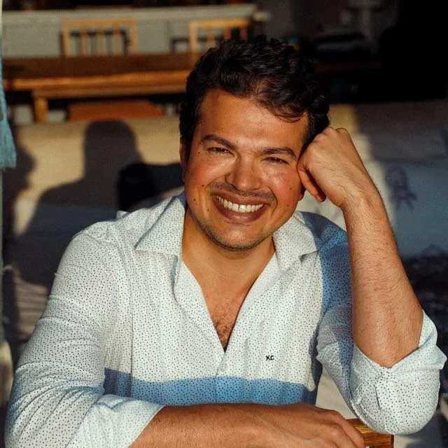A smiling man with dark hair, wearing a white shirt with small dark dots, sitting outdoors with a blurred background of outdoor furniture.