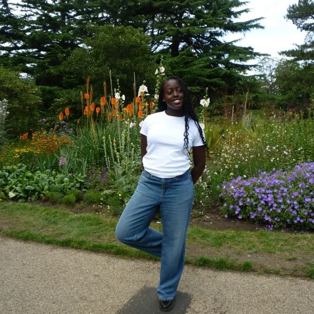 A young woman smiling and posing in a garden with colorful flowers and tall green trees in the background.