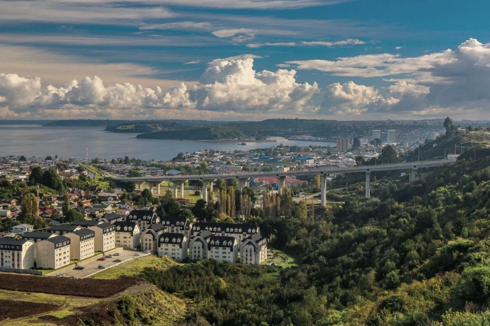 Vista panorámica de la ciudad de Puerto Montt con edificios, un puente, el mar y colinas verdes, bajo un cielo con nubes.