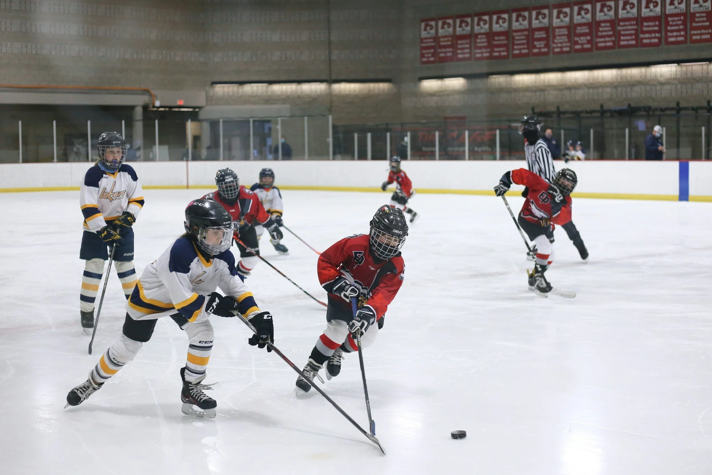 Youth hockey players wearing helmets and jerseys playing ice hockey on an indoor rink, with a referee in a striped shirt watching in the background.