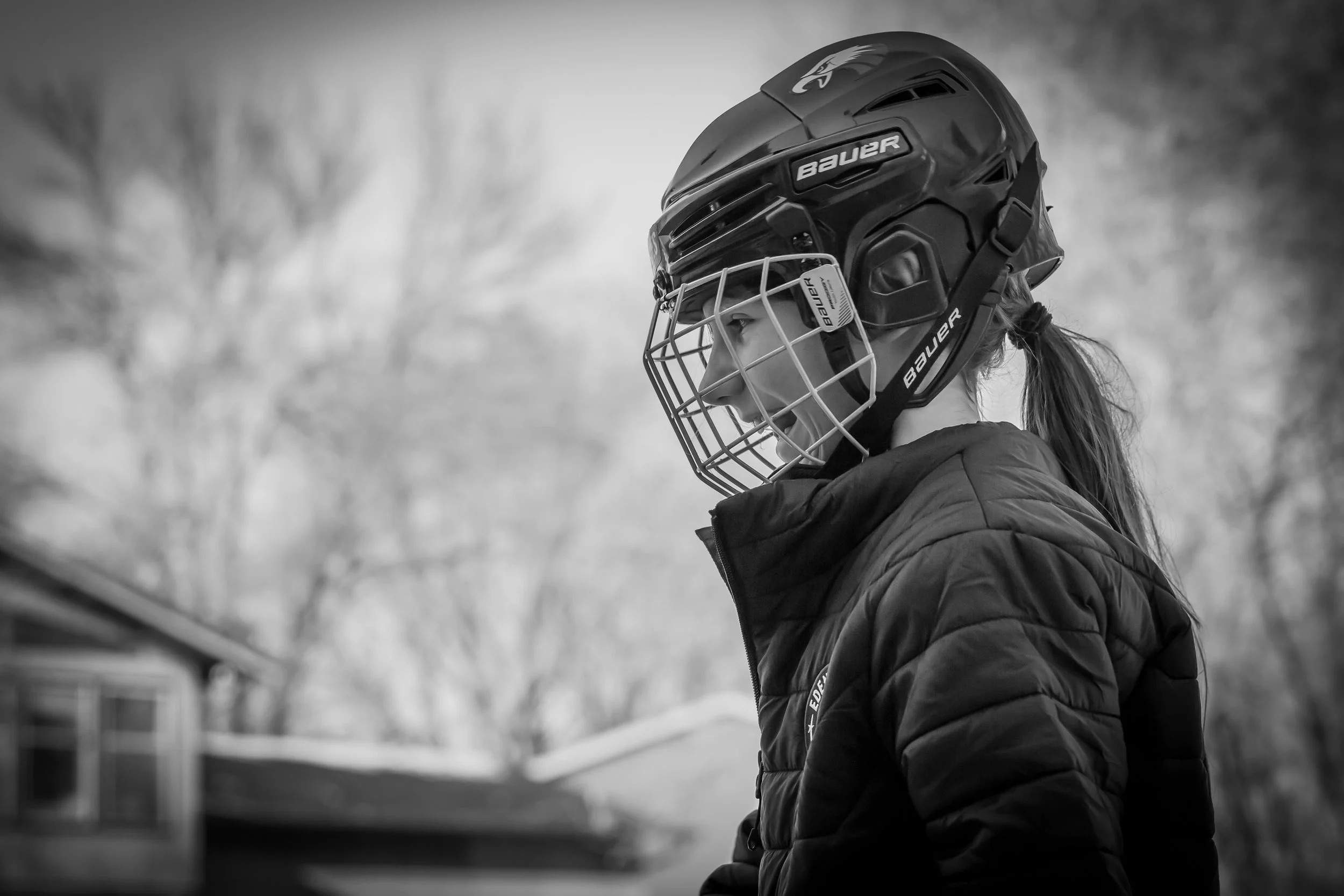 Side profile of a female hockey player wearing a Bauer helmet and face cage, dressed in a padded jacket, outdoors in winter. Background shows trees and houses in black and white.