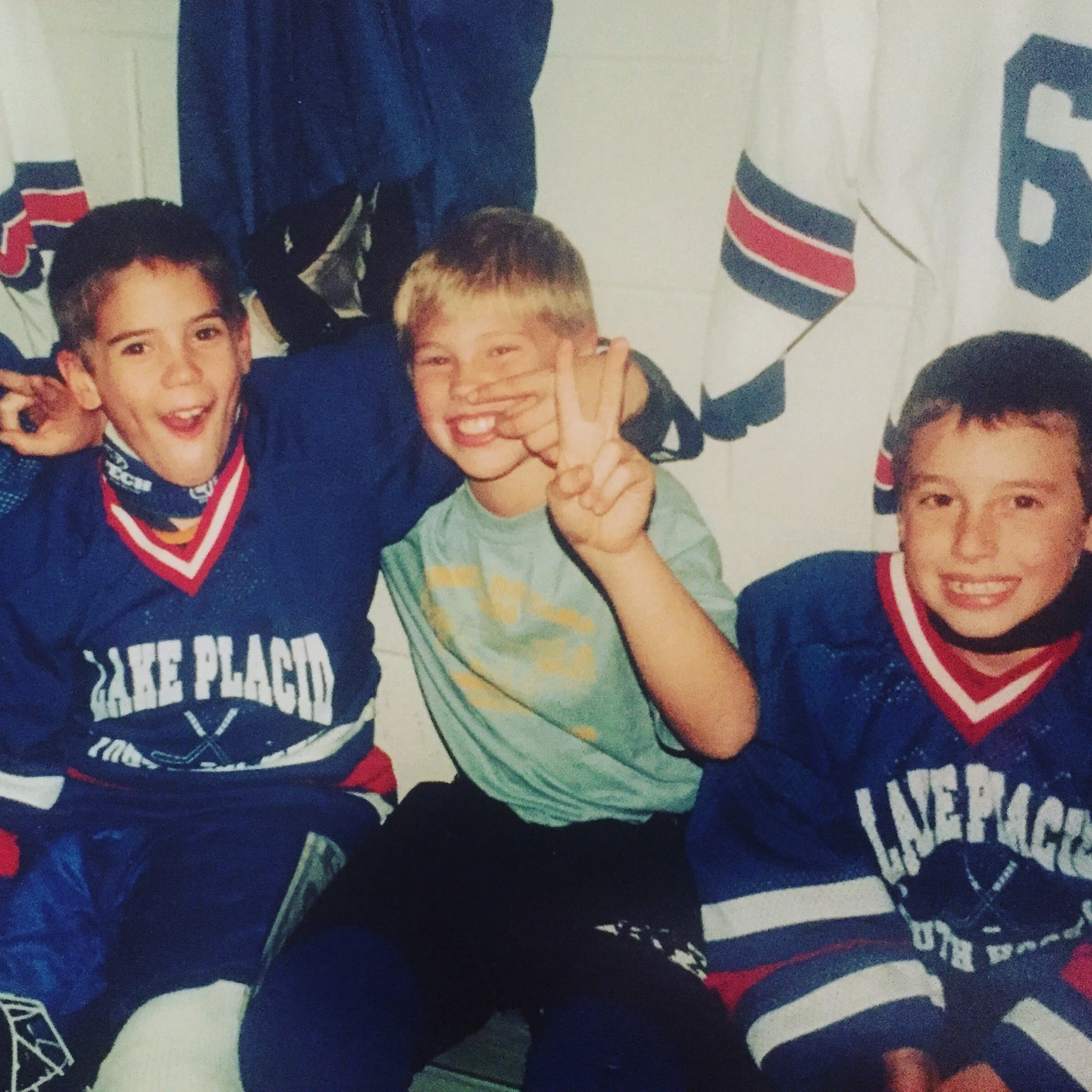 Two young boys smiling and posing for a photo, with one making a peace sign. Behind them, there are hockey jerseys and American flags hanging on the wall, suggesting a sports or team celebration.