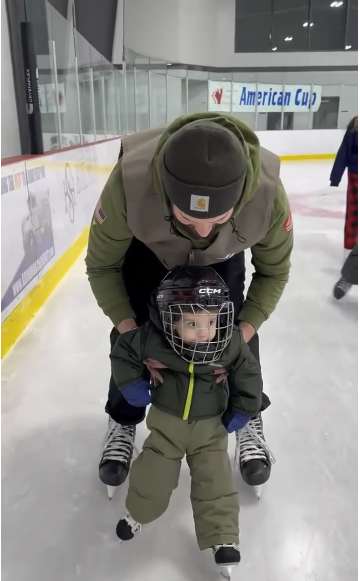 An adult helping a young child on an ice rink, with the child wearing a helmet and skating gear. The adult is bent over supporting the child, both on ice skates. In the background, an American Cup banner is visible.