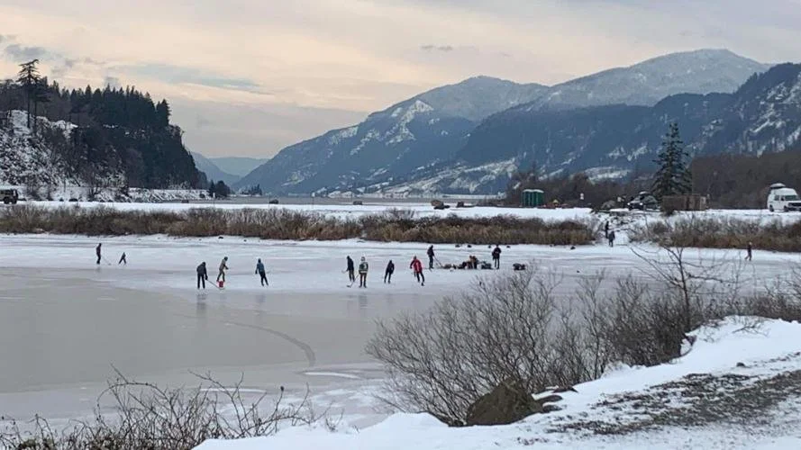 People ice skating on a frozen lake surrounded by snow-covered landscape and mountains in the background.