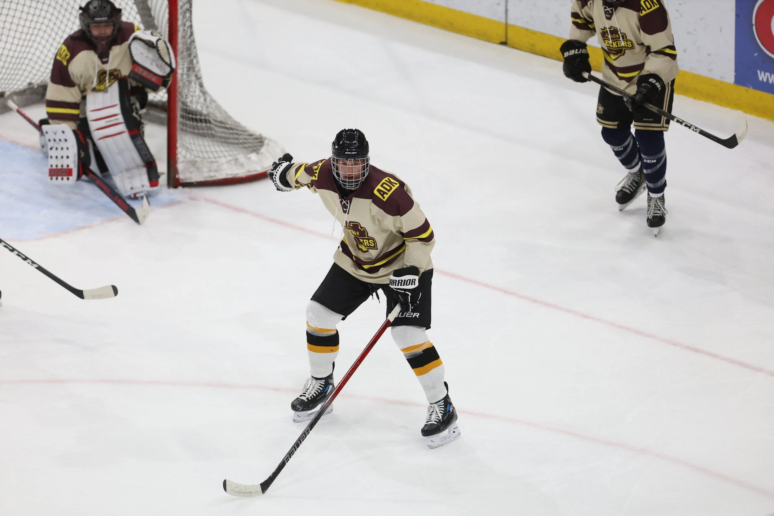 Ice hockey game scene with a player in maroon and beige uniform skating on ice, another player near the goal, and a goalie in maroon and beige gear standing in front of the net.