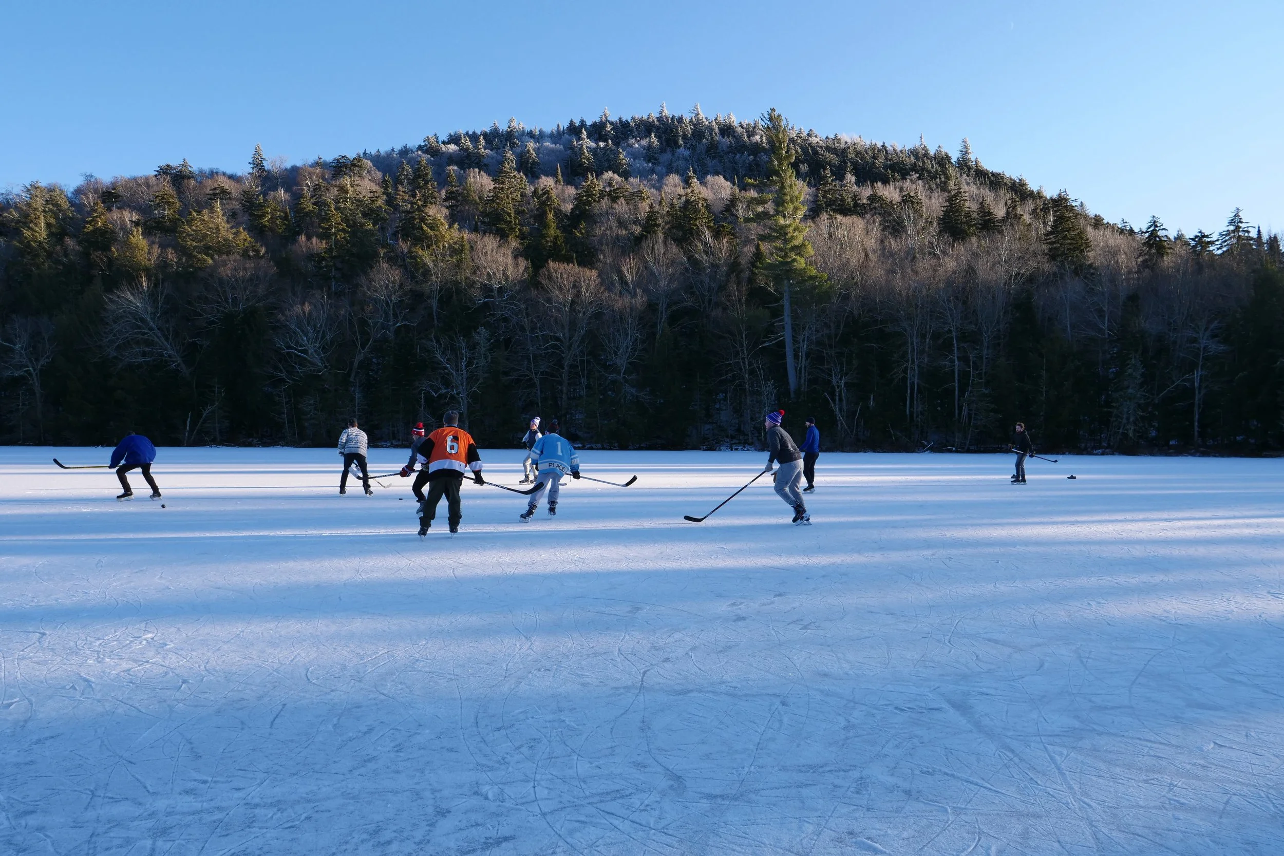 People skating on an outdoor ice rink with a forest of pine trees and a mountain in the background under a clear blue sky.