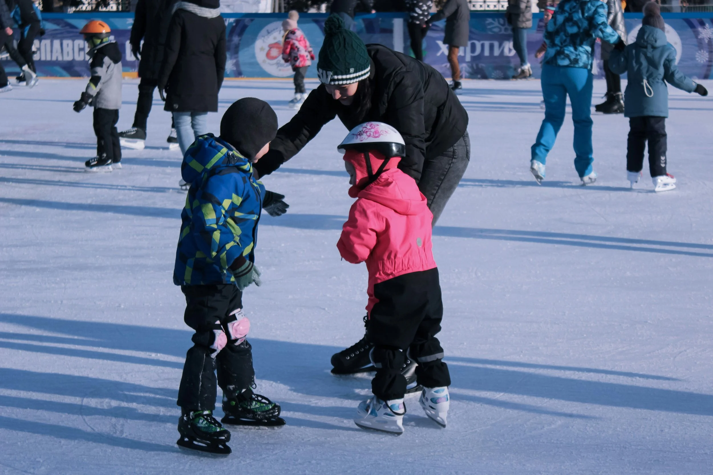 People ice skating outdoors on a sunny day, including children and adults wearing winter clothing and skates.